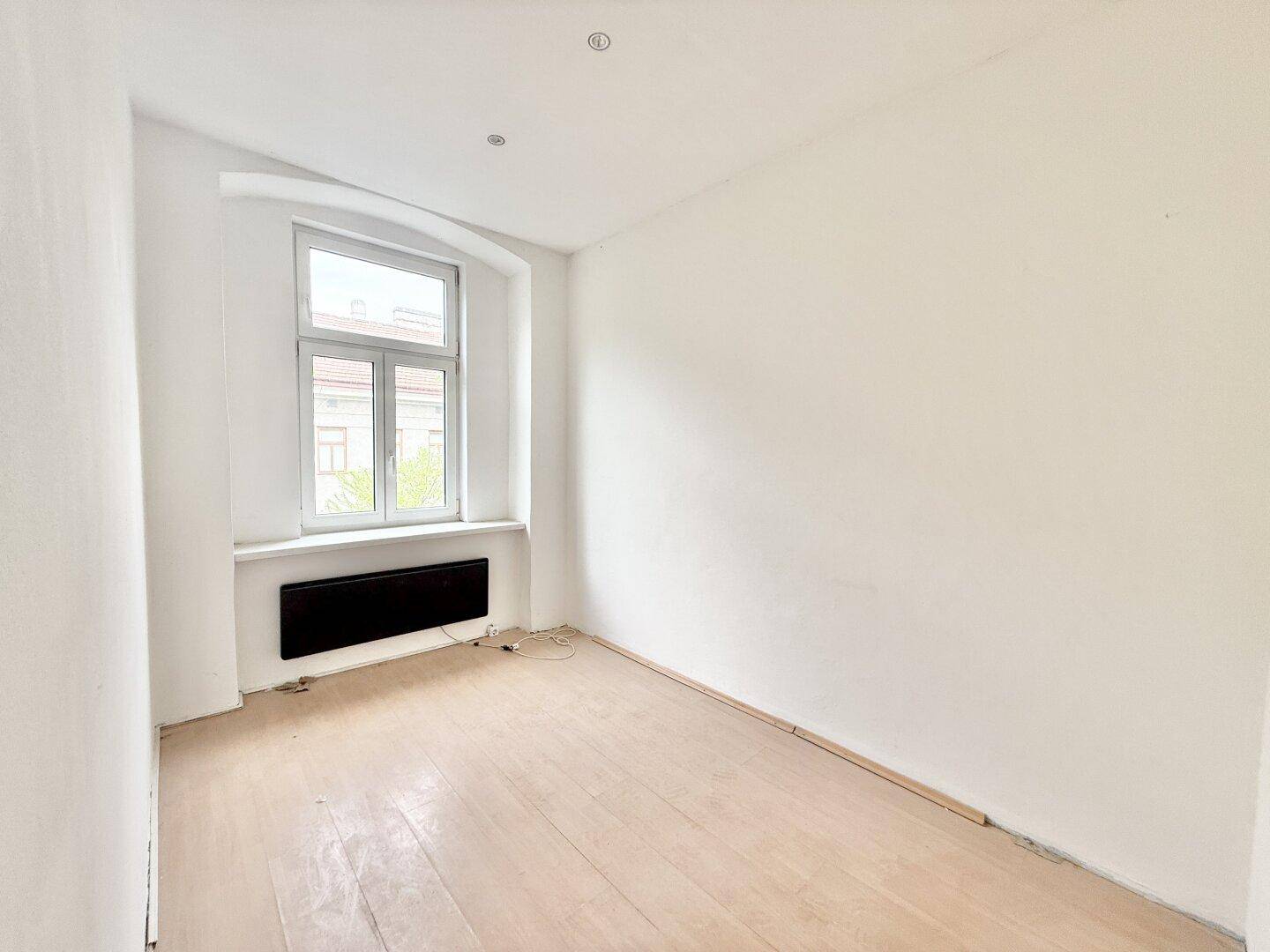 Leerer Raum mit hellem Holzboden, großem Fenster und Heizkörper, bereit zur Gestaltung.