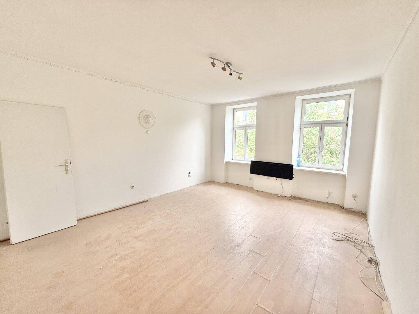 Geräumiger leerer Raum mit zwei großen Fenstern, Holzboden und Stuckdecke.