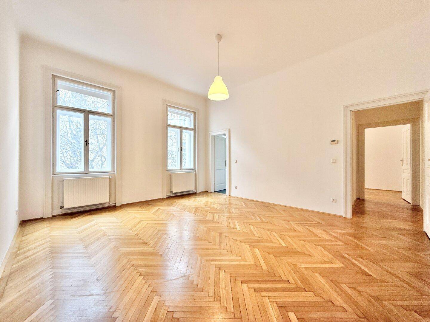 Geräumiges, leeres Wohnzimmer mit klassischem Fischgrätparkett, hohen Decken und großen Fenstern.