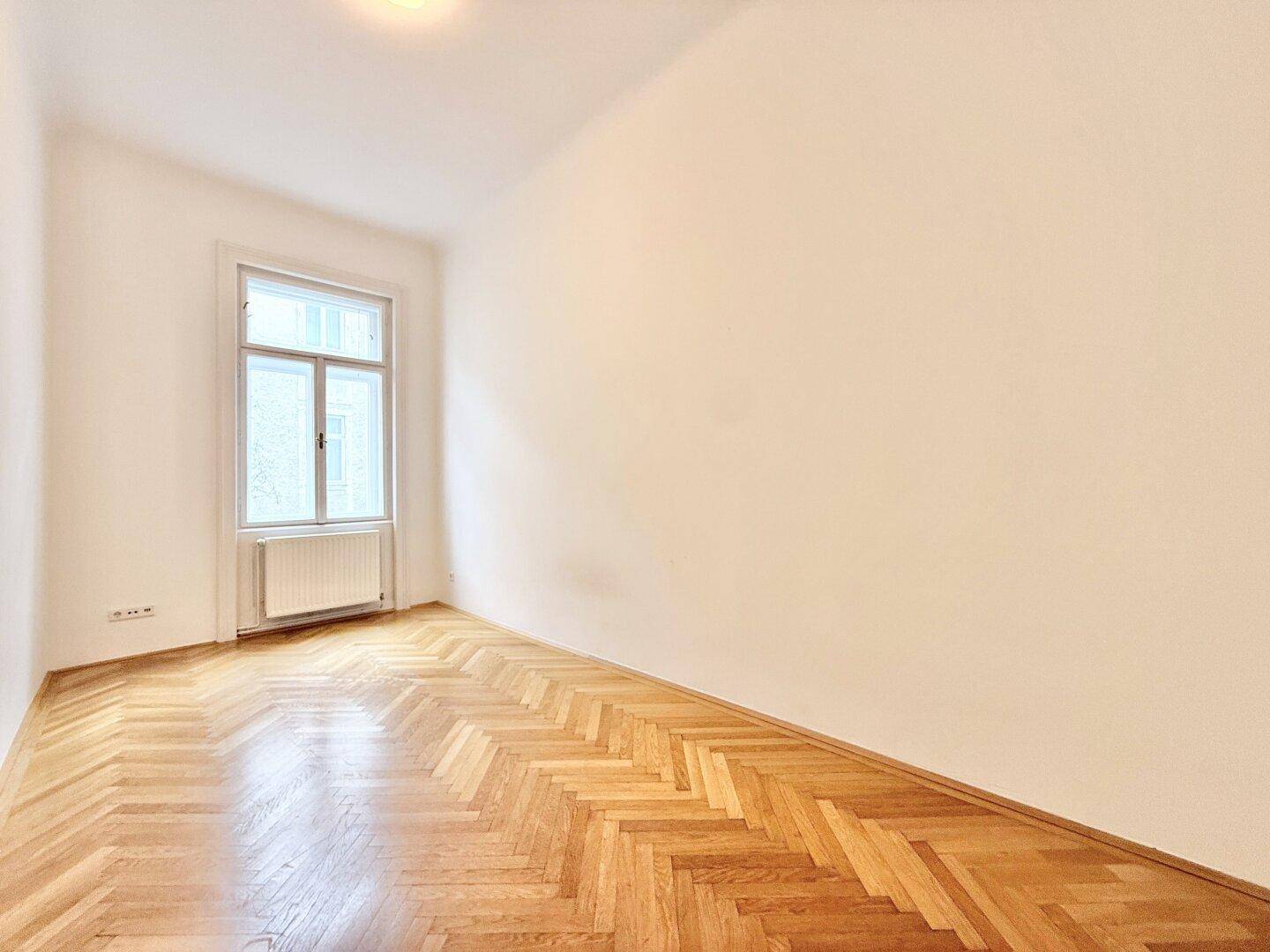 Helles, leeres Zimmer mit Fischgrätparkett und einem Fenster, ideal als Schlaf- oder Arbeitszimmer.