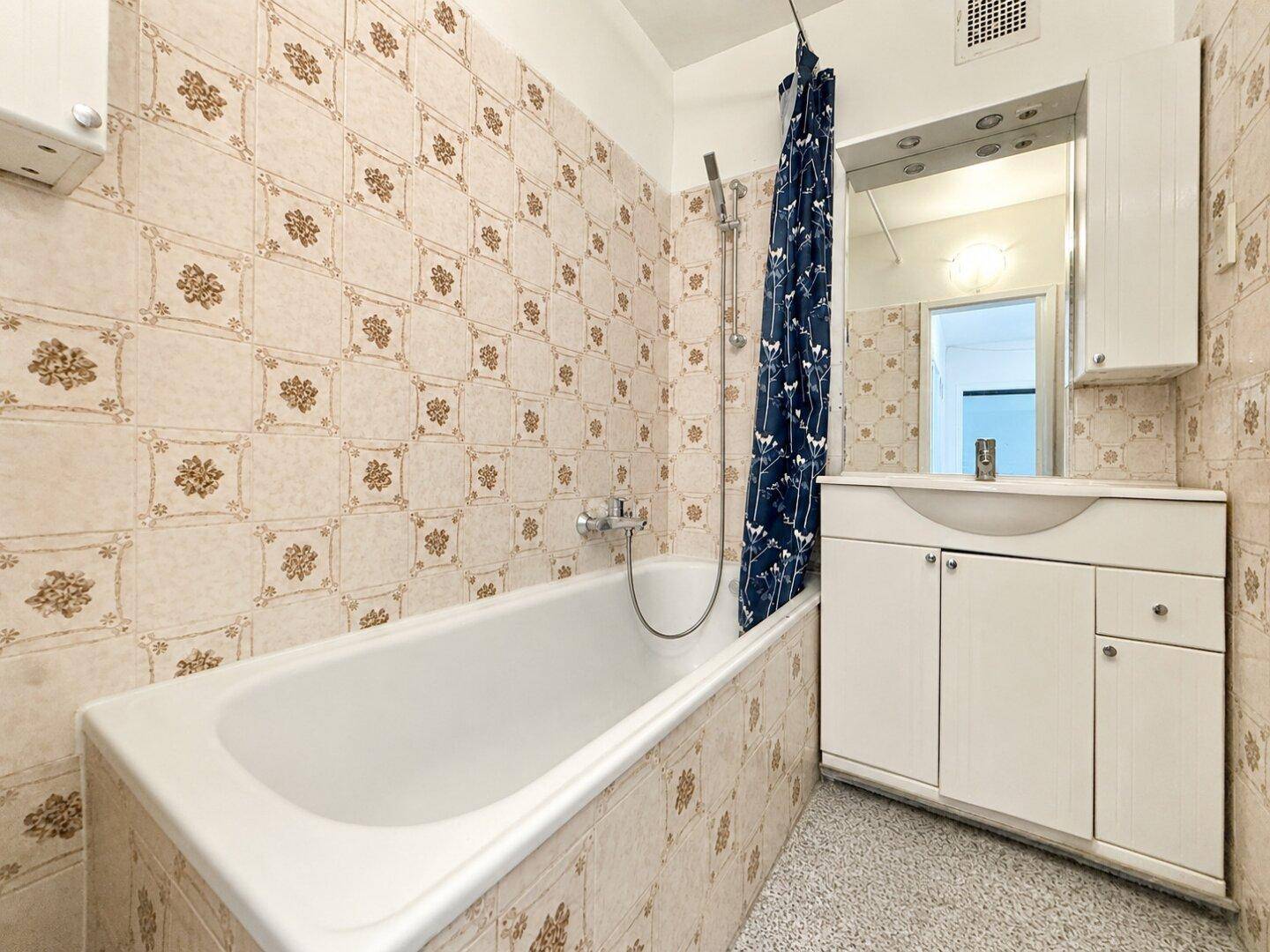 Älteres Badezimmer mit Badewanne, Duschvorhang und gemusterten Fliesen.
