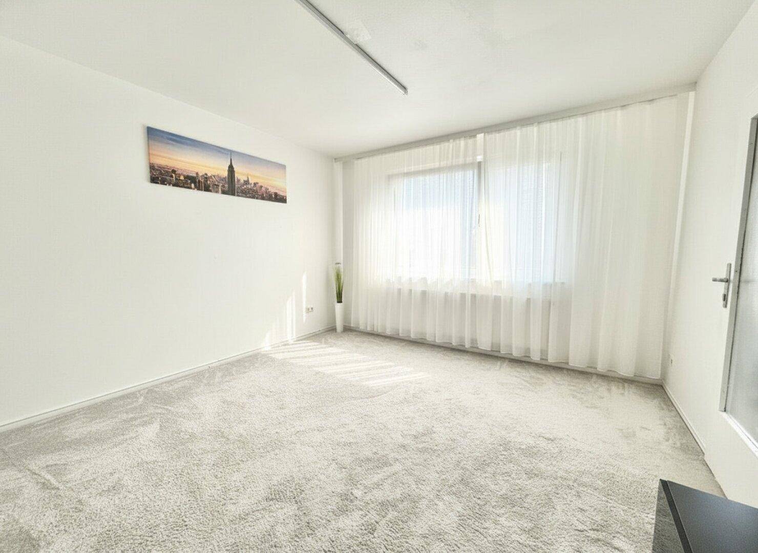 Geräumiges, unmöbliertes Wohnzimmer mit hellem Teppichboden und großem Fenster.
