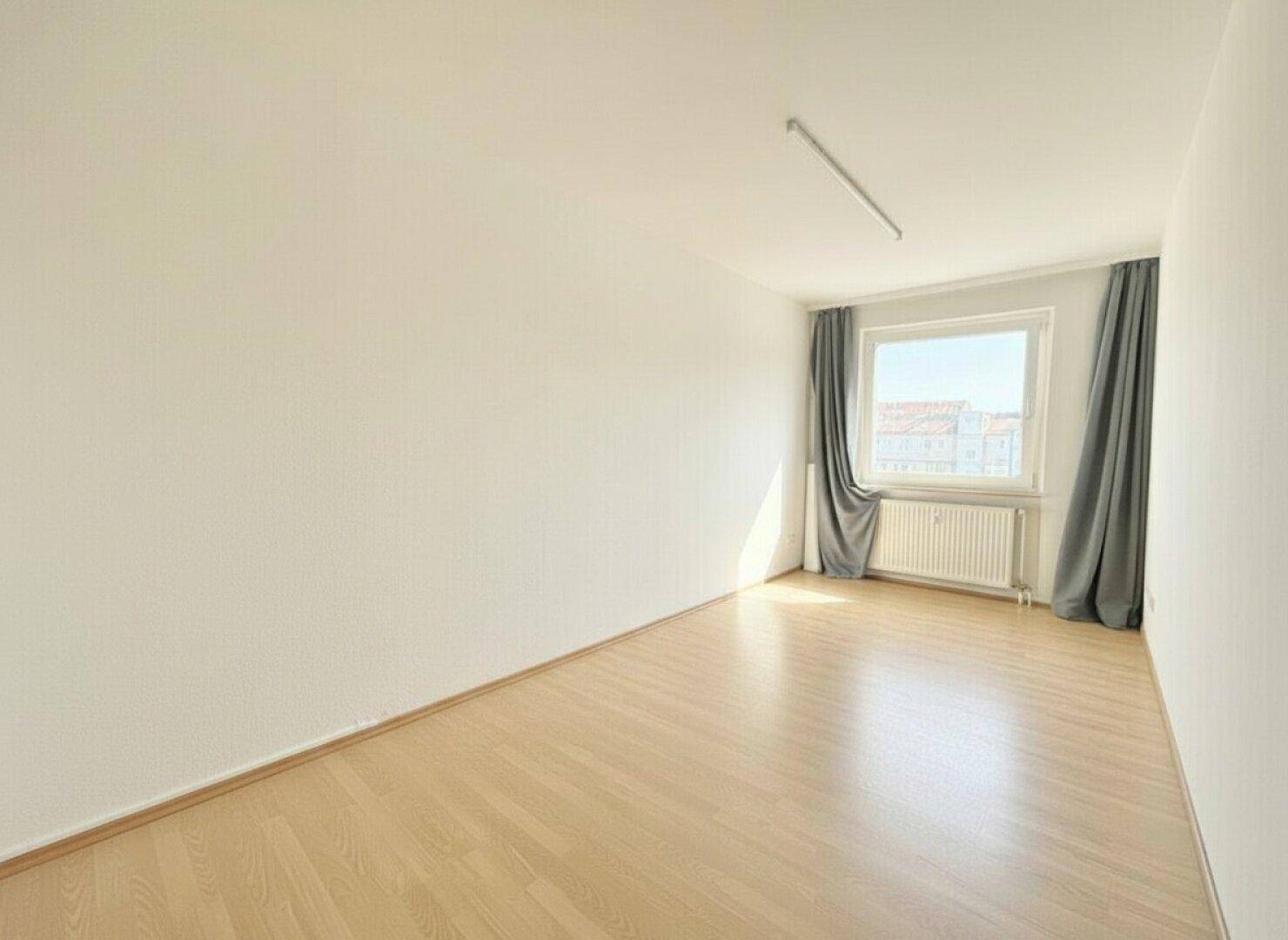 Leeres Schlafzimmer mit hellem Parkettboden, Fenster und Heizkörper unter dem Fenster.