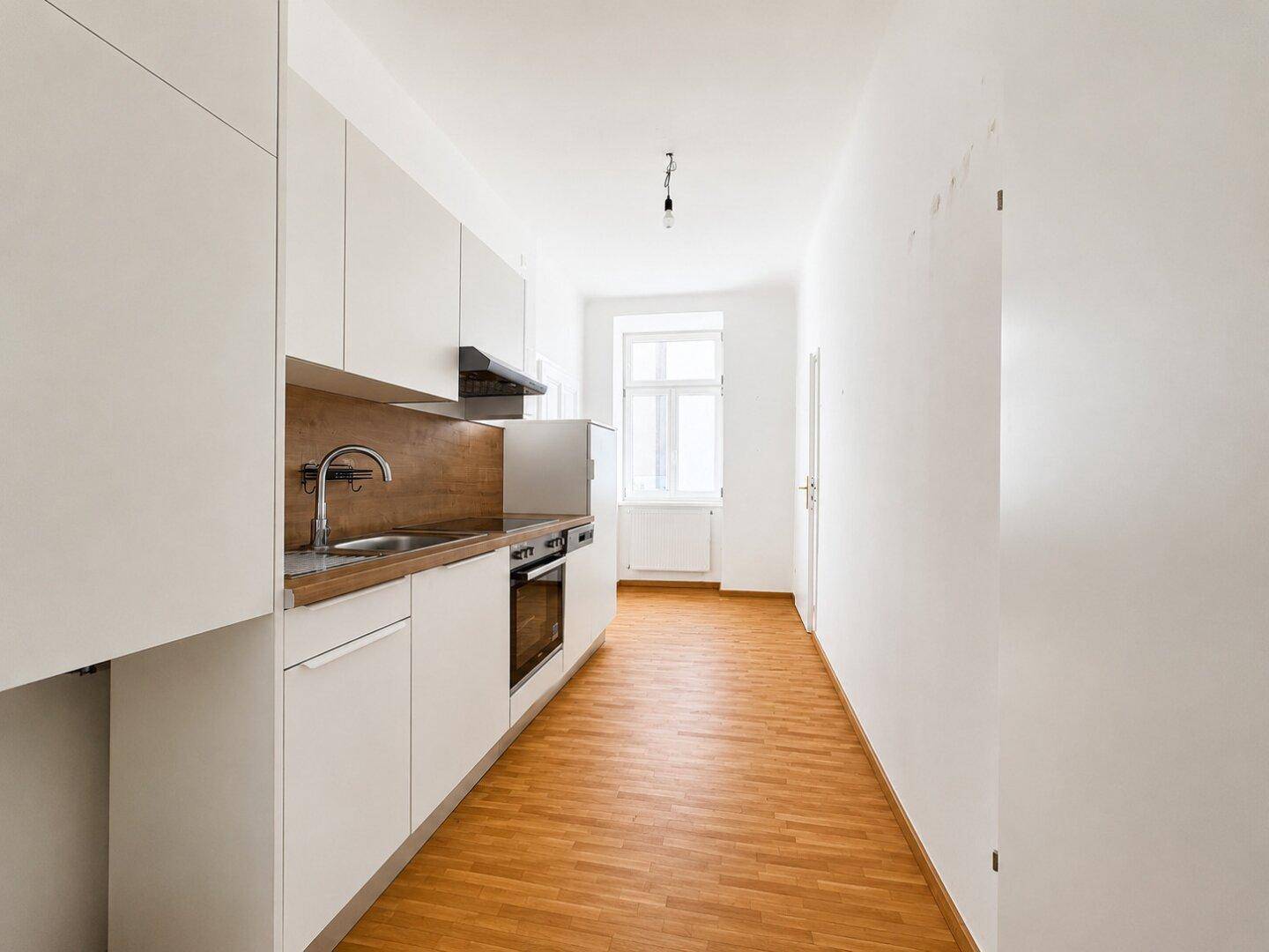 Schmale, helle Küche mit weißer Einbauküche, Holzarbeitsplatte und Fenster am Ende des Raumes.