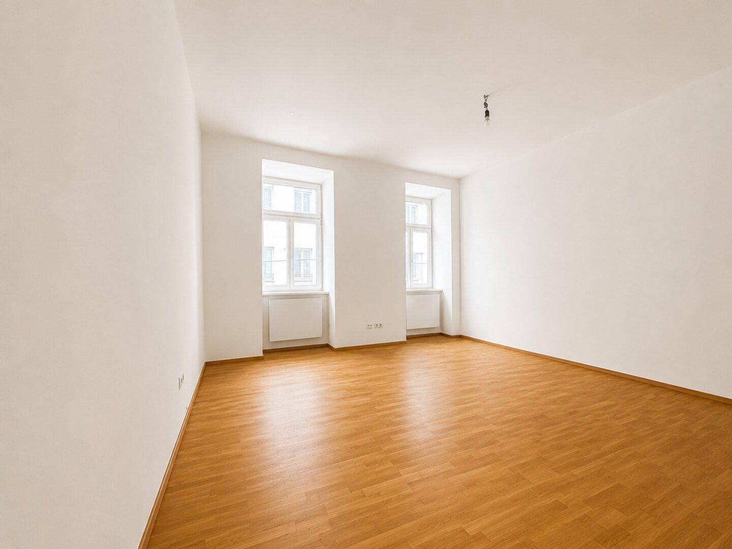 Helles, leeres Zimmer mit Holzboden und zwei großen Fenstern, bereit zur individuellen Gestaltung.