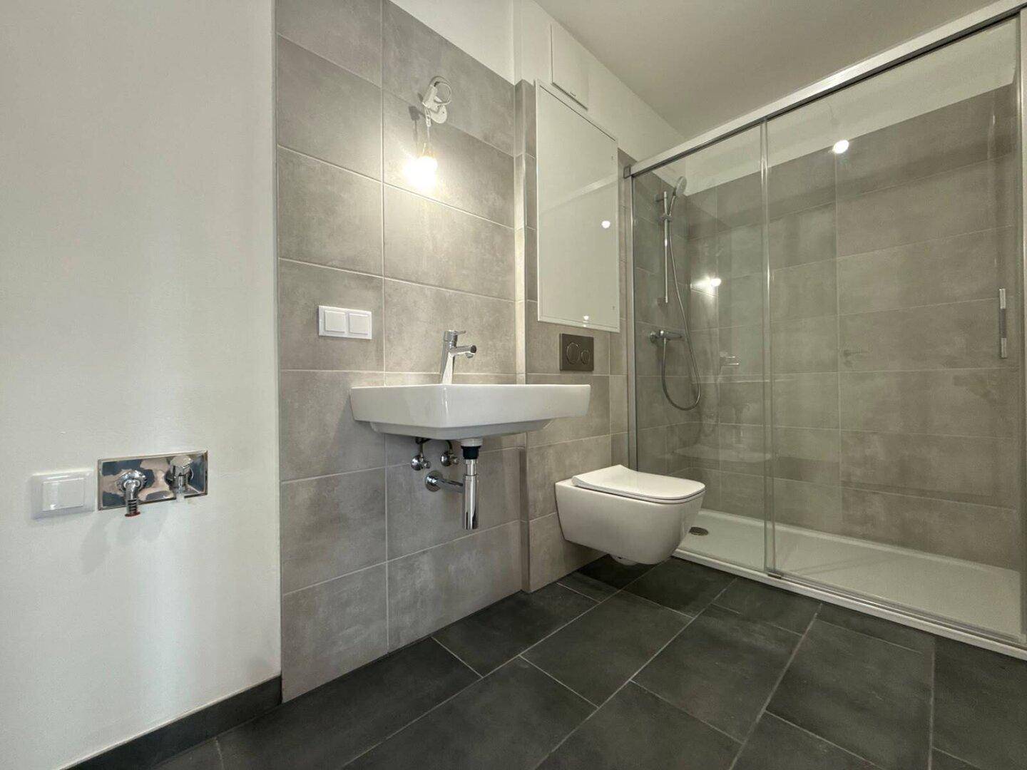 Modernes Badezimmer mit grauen Fliesen, einer geräumigen Dusche, Waschbecken und wandhängendem WC.