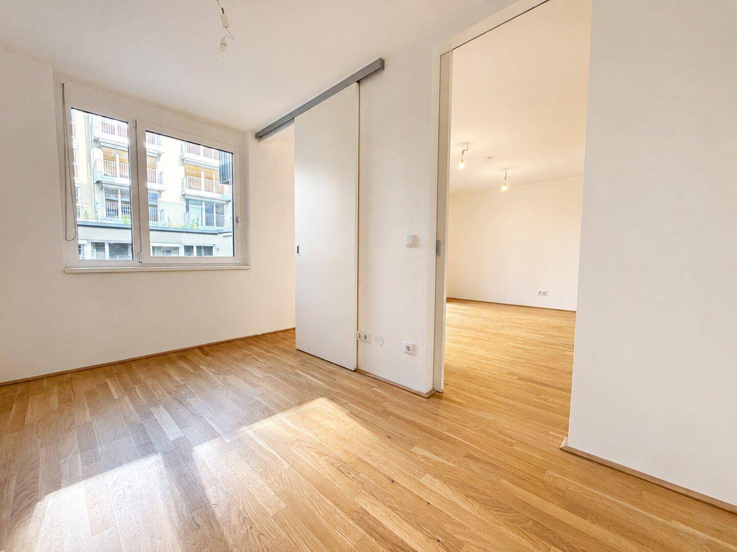 Leeres Zimmer mit Holzboden, großem Fenster und einer praktischen Schiebetür zu einem weiteren Raum.