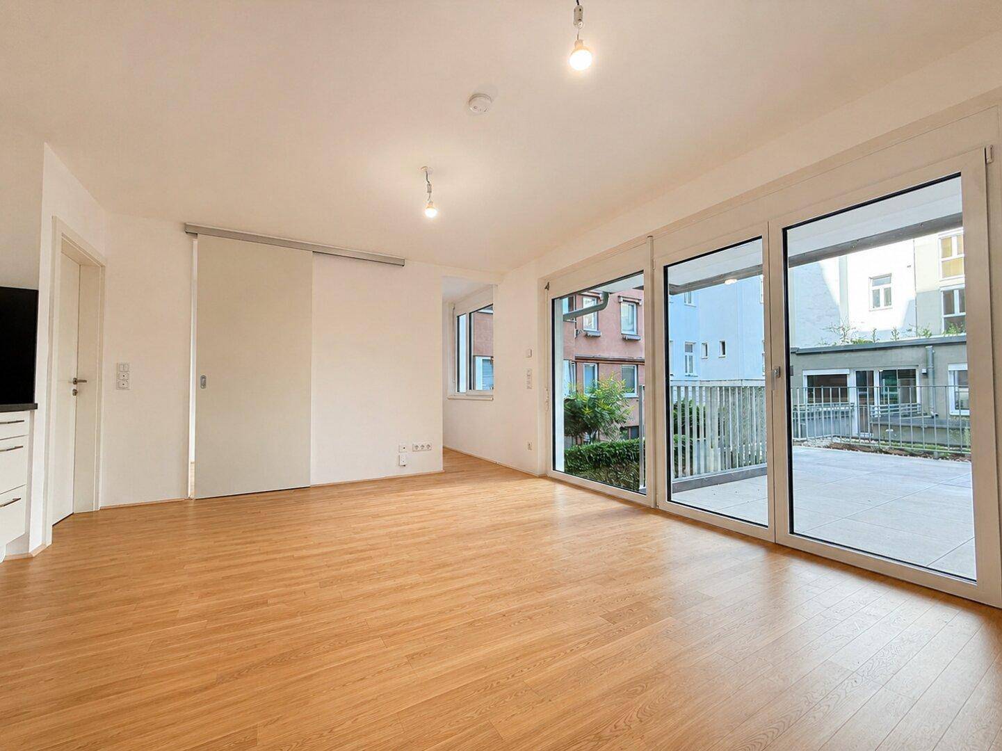 Geräumiger, leerer Wohnbereich mit Holzboden und großen Fenstern, die zur Terrasse führen.