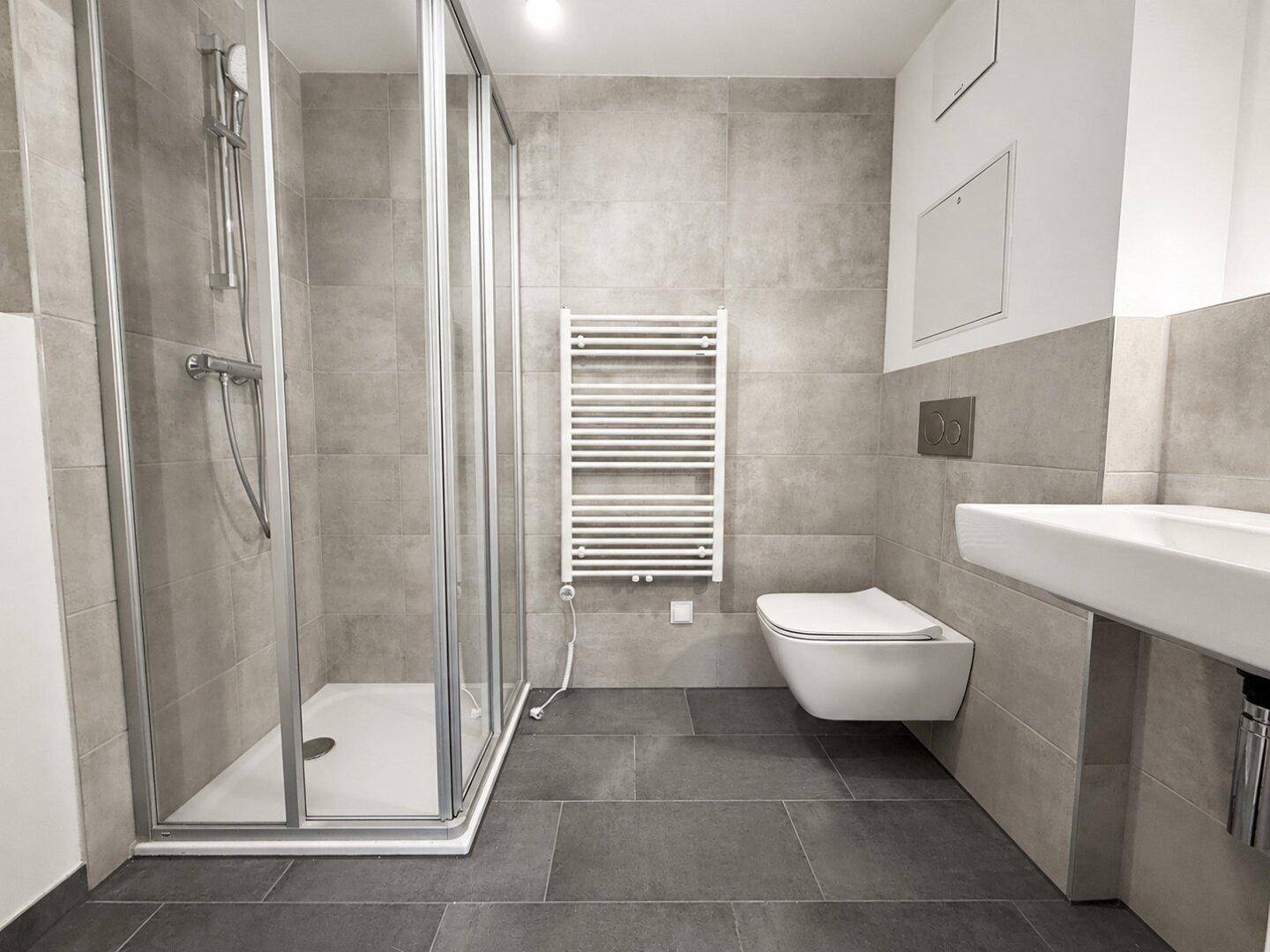 Modernes Badezimmer mit grauen Fliesen, Duschkabine, wandhängendem WC und Handtuchheizkörper.