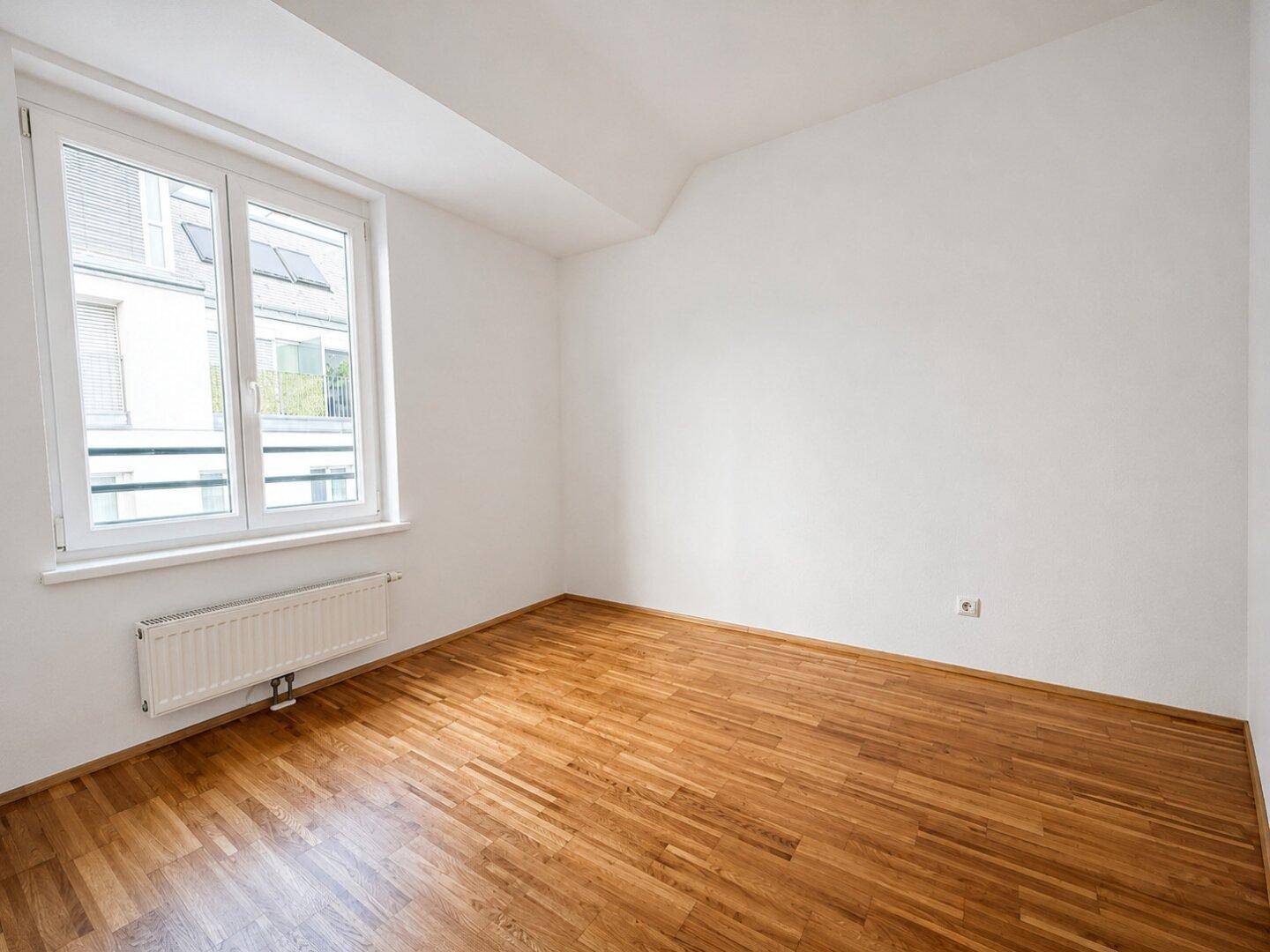 Heller, leerer Raum mit Holzboden und großem Fenster, ideal als Kinder- oder Arbeitszimmer.