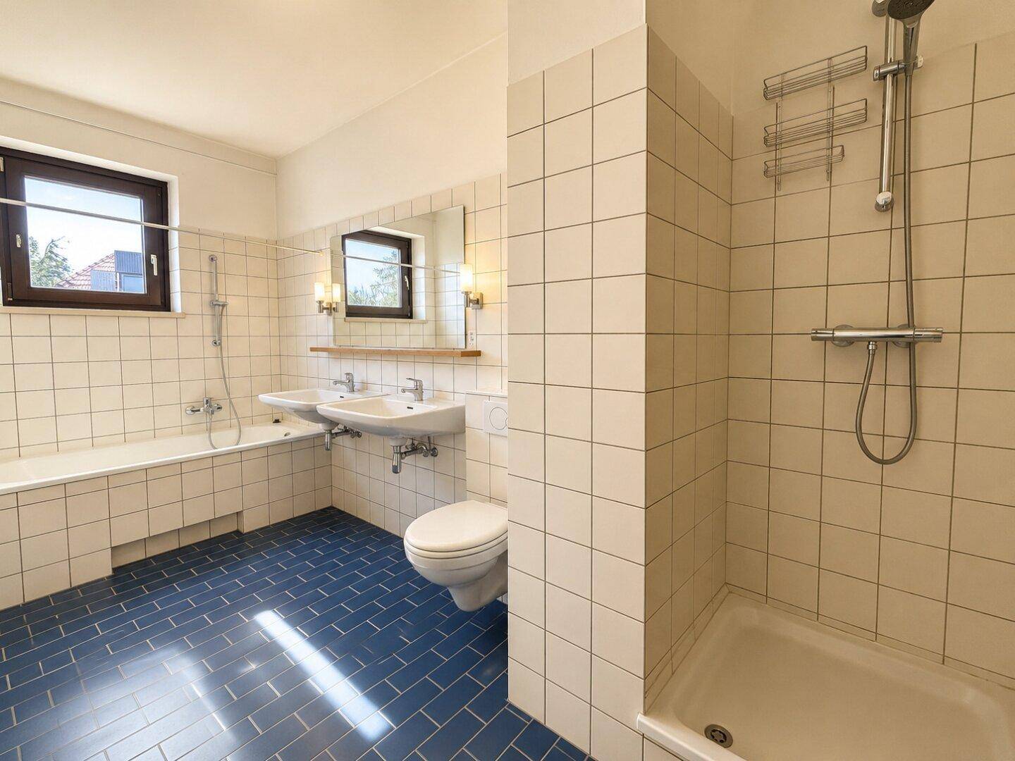 Badezimmer mit blauen Bodenfliesen, Doppelwaschbecken, Badewanne und Duschbereich.