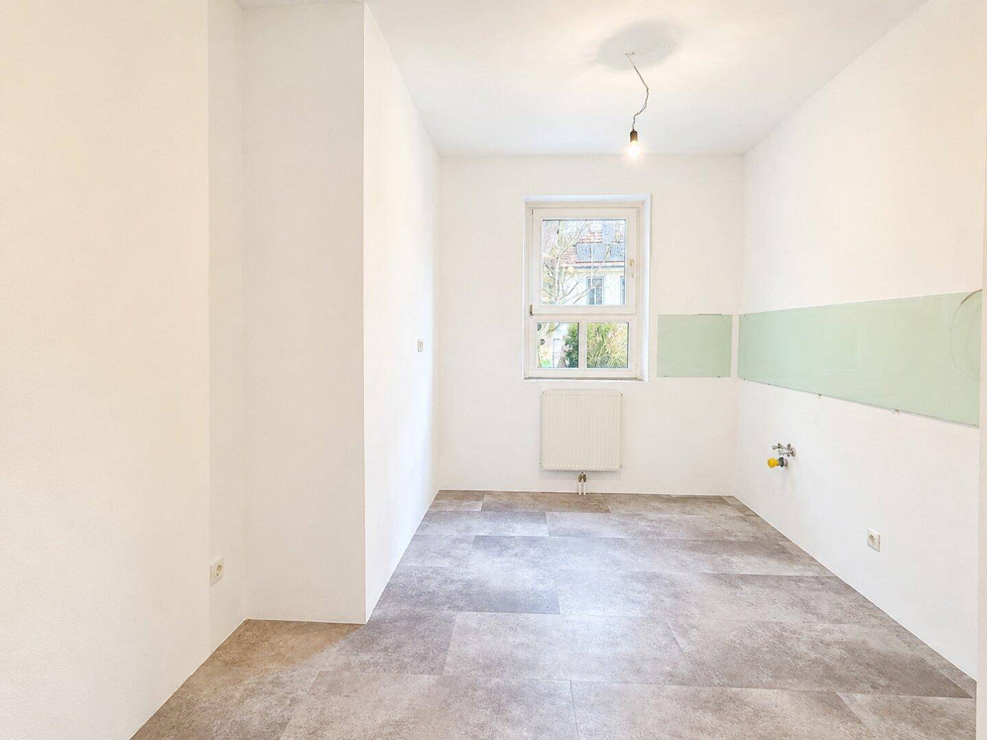 Leerer Küchenraum mit Fliesenboden, Fenster und Anschlüssen für eine Einbauküche.