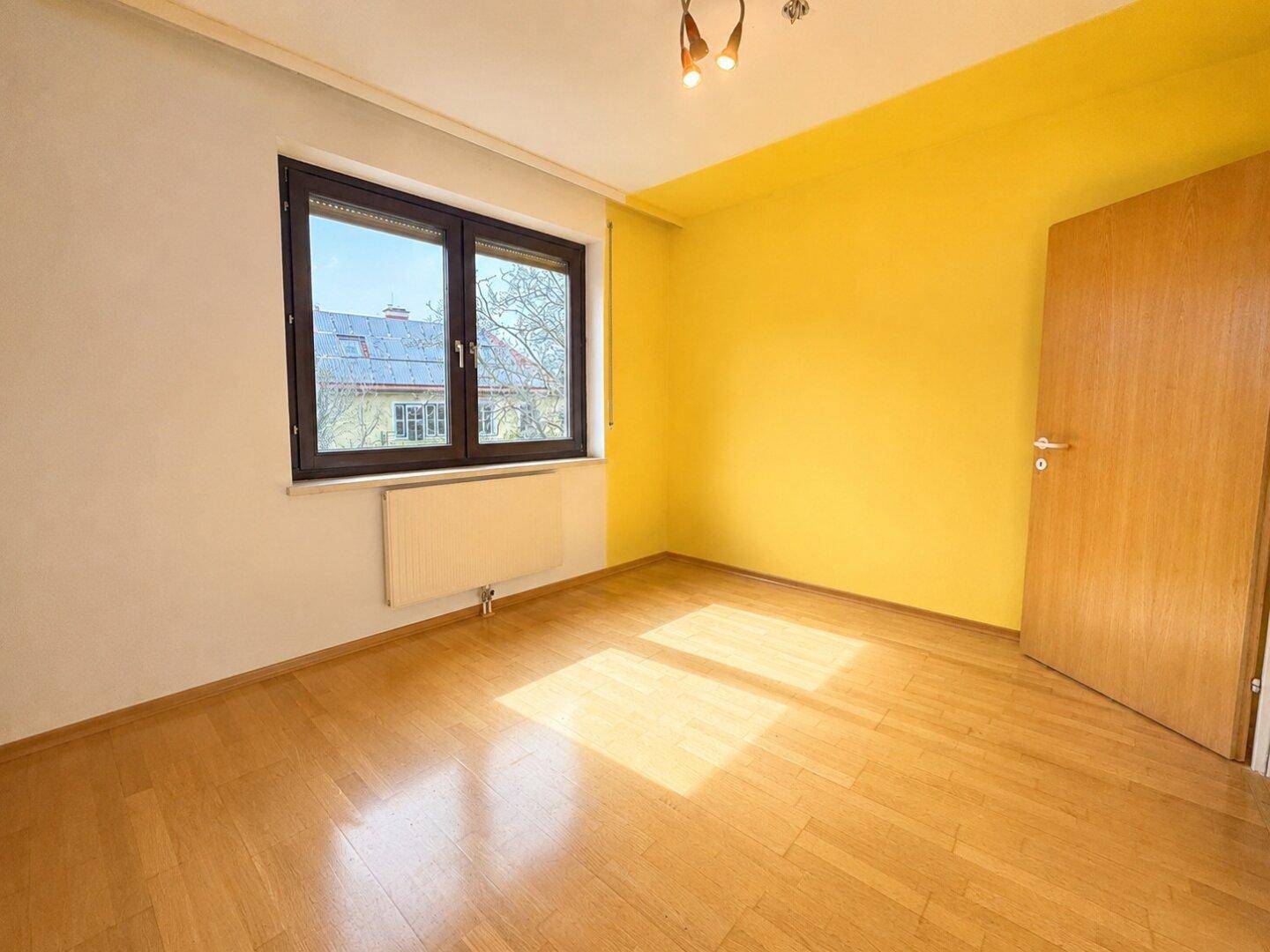 Helles Schlafzimmer mit Parkettboden, gelber Akzentwand und großem Fenster.