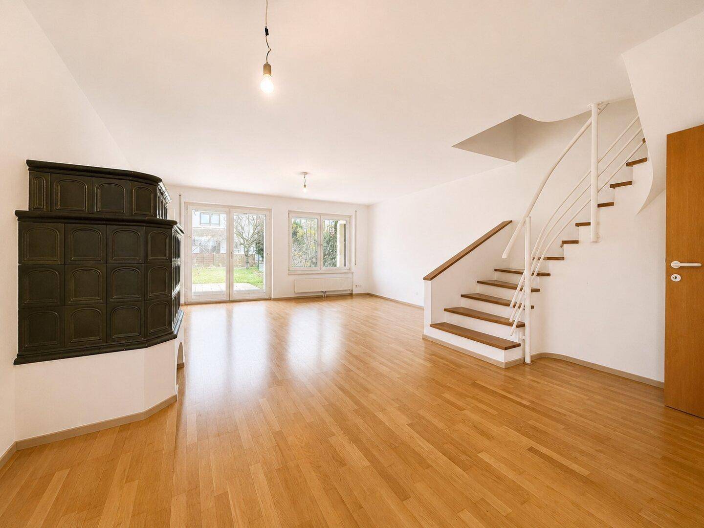 Geräumiges Wohnzimmer mit hellem Parkettboden, Kachelofen und Treppe zum Obergeschoss.