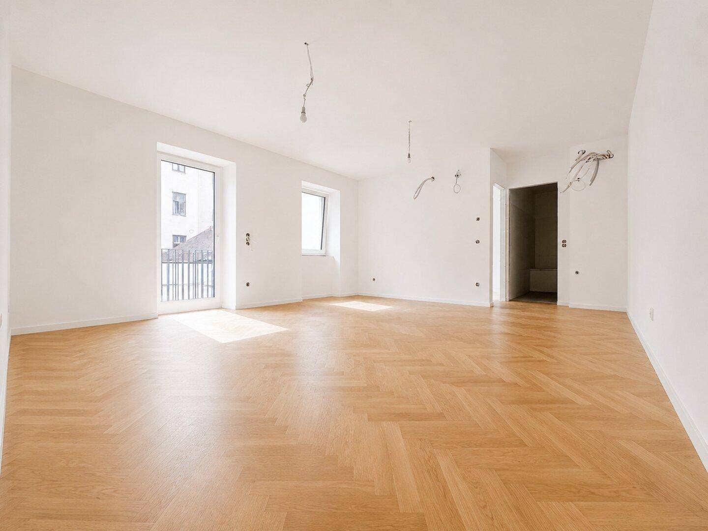 Geräumiges, leeres Zimmer mit Fischgrätparkett und Zugang zu einem französischen Balkon.