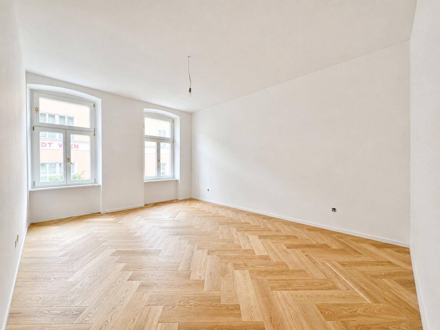 Heller, leerer Raum mit elegantem Fischgrätparkett und zwei großen Fenstern.