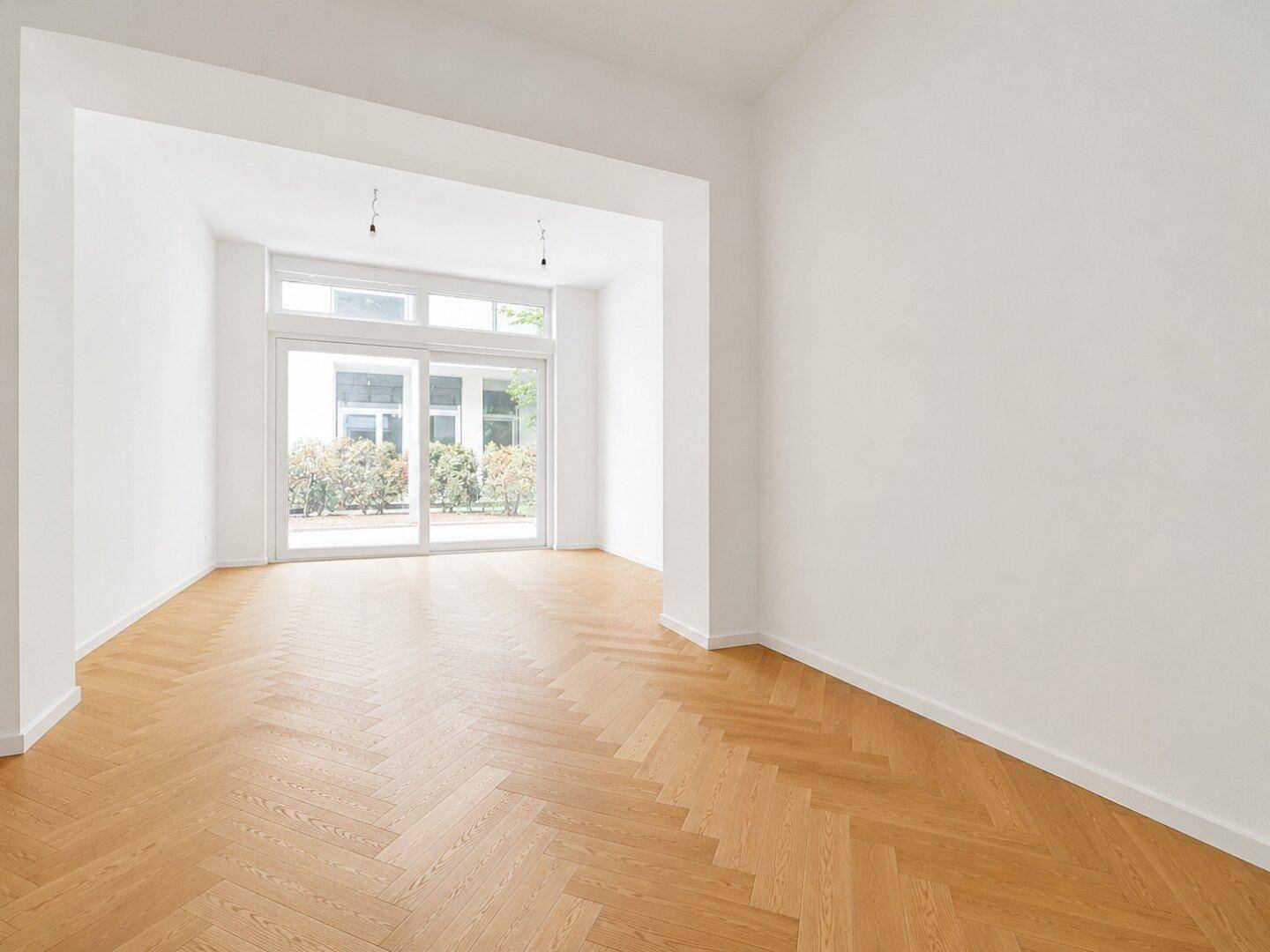 Geräumiger, heller Raum mit Fischgrätparkett und großen Fenstern, die viel Tageslicht hereinlassen.