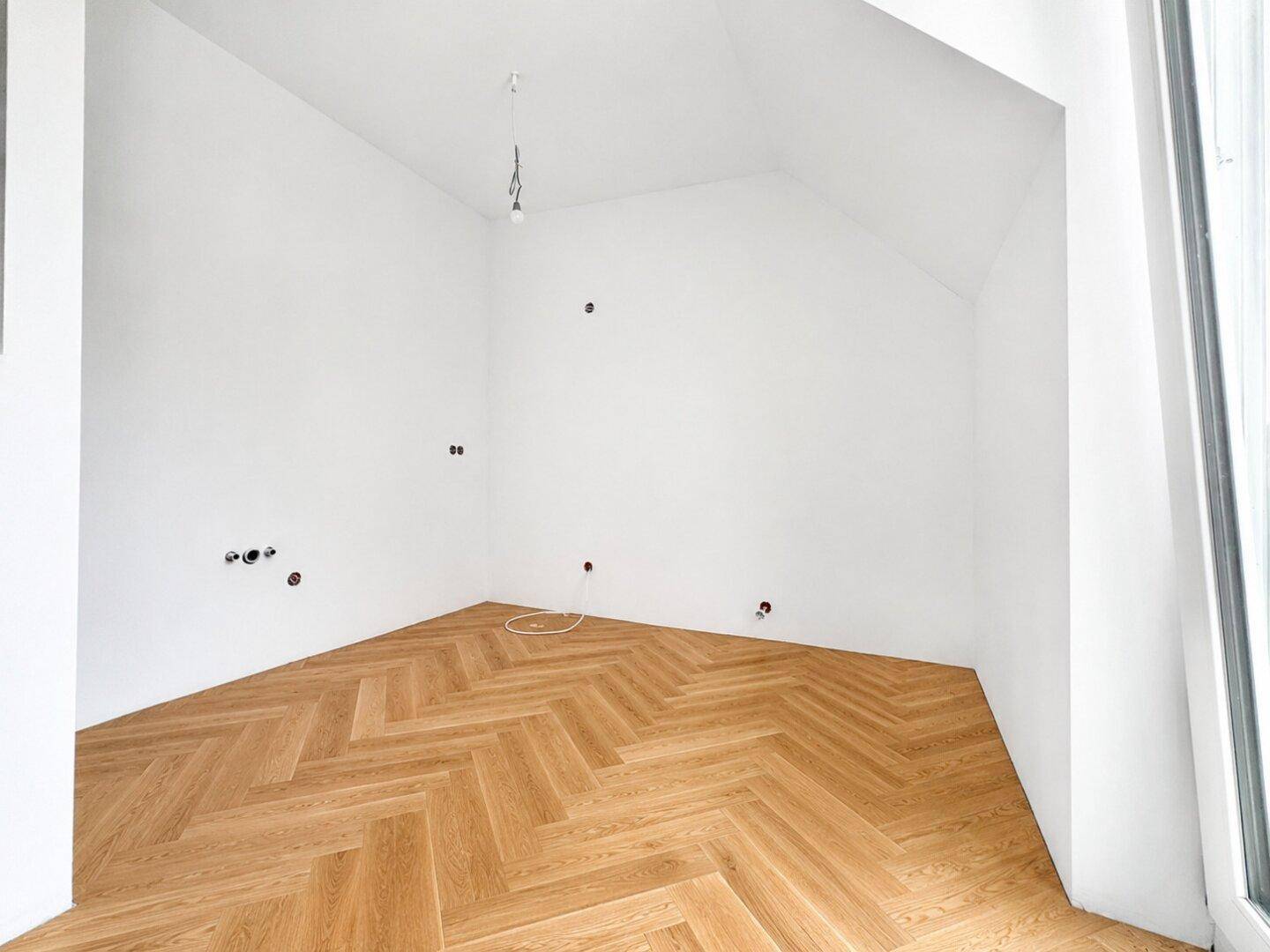 Leerer Raum mit Fischgrätparkett, schrägen Wänden und sichtbaren Anschlüssen.