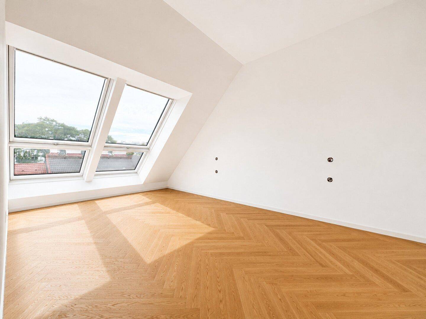Lichtdurchfluteter Raum mit Fischgrätparkett, großen Dachfenstern und schrägen Wänden.