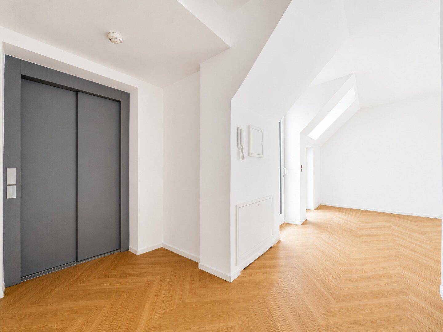 Heller Flur mit Fischgrätparkett und modernem Aufzug, der alle Etagen erschließt.