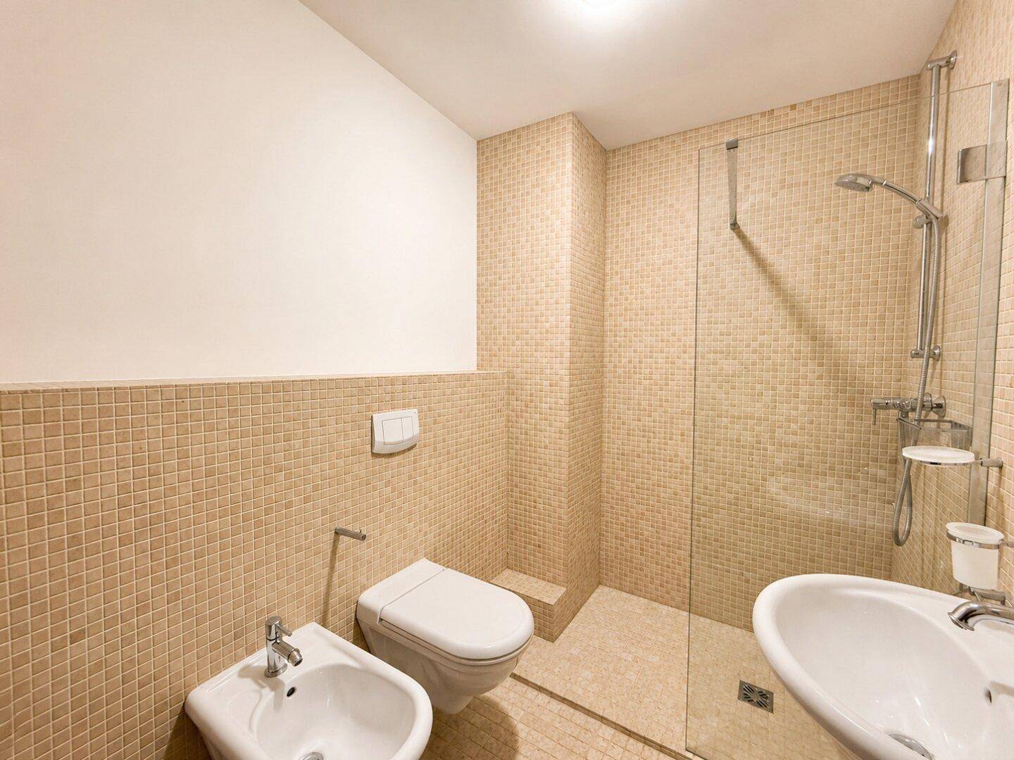 Badezimmer mit beigen Mosaikfliesen, Dusche, WC, Bidet und Einzelwaschbecken.