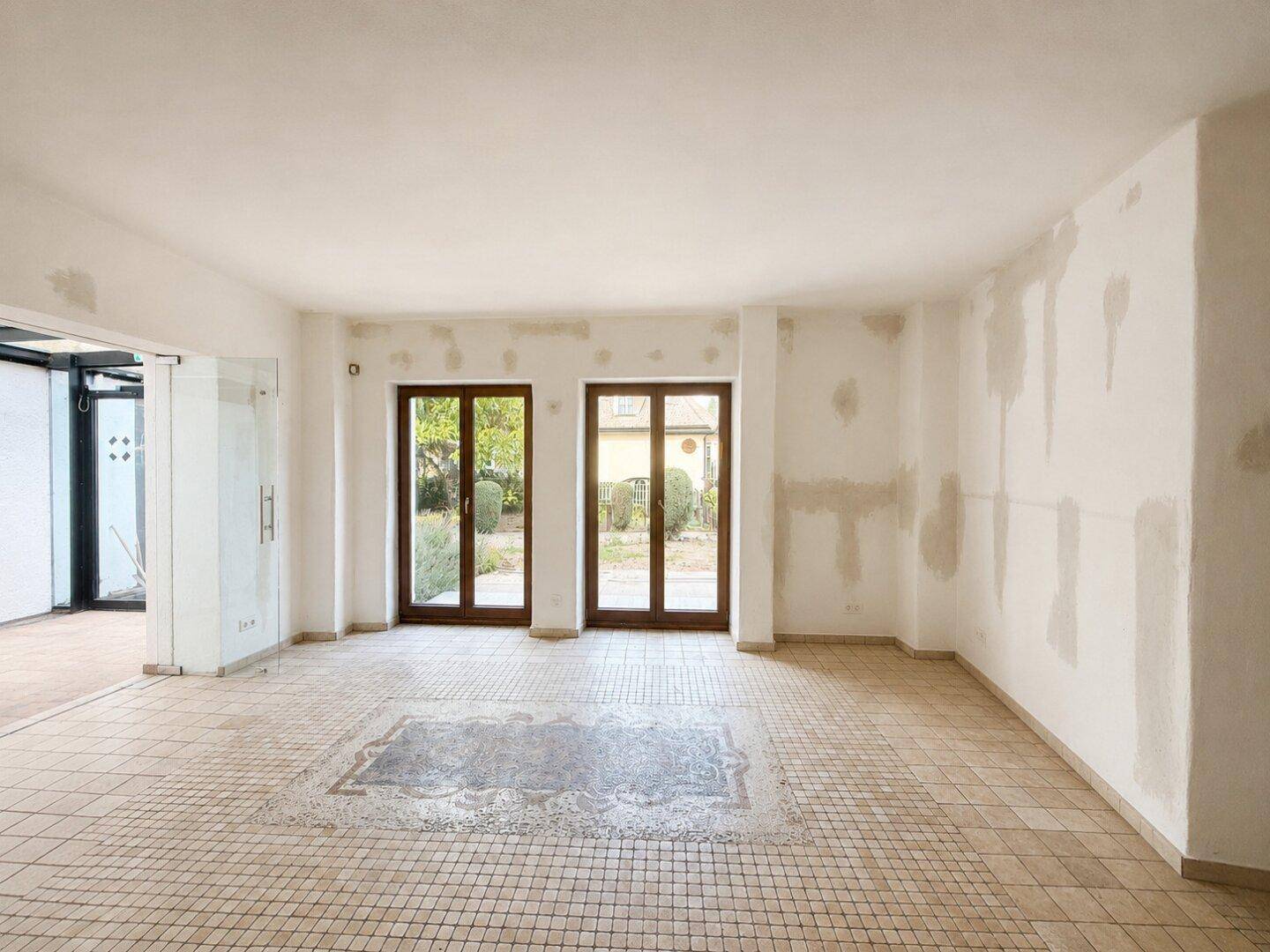 Geräumiger Raum im Rohzustand mit Fliesenboden und zwei Fenstertüren zum Garten.