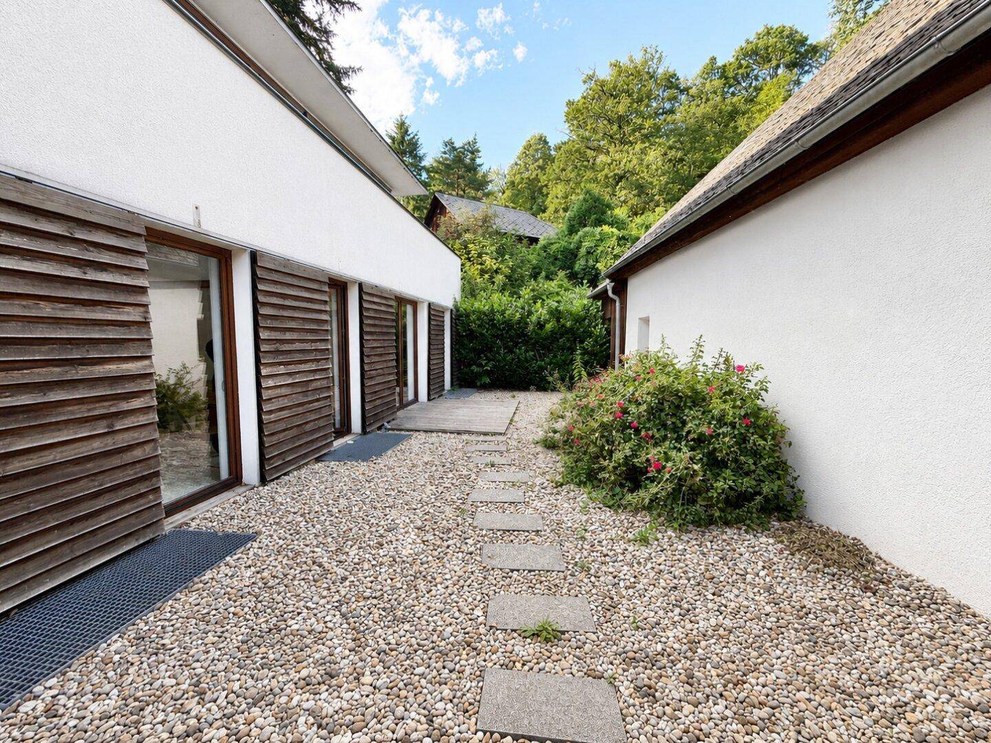 Kiesweg zwischen zwei Gebäudeteilen mit Holzfassade und grüner Bepflanzung.