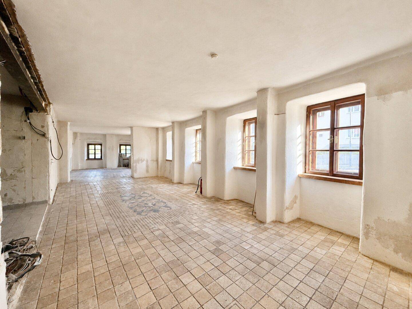 Langer, heller Raum mit Fliesenboden und mehreren Fenstern, bereit für die Renovierung.