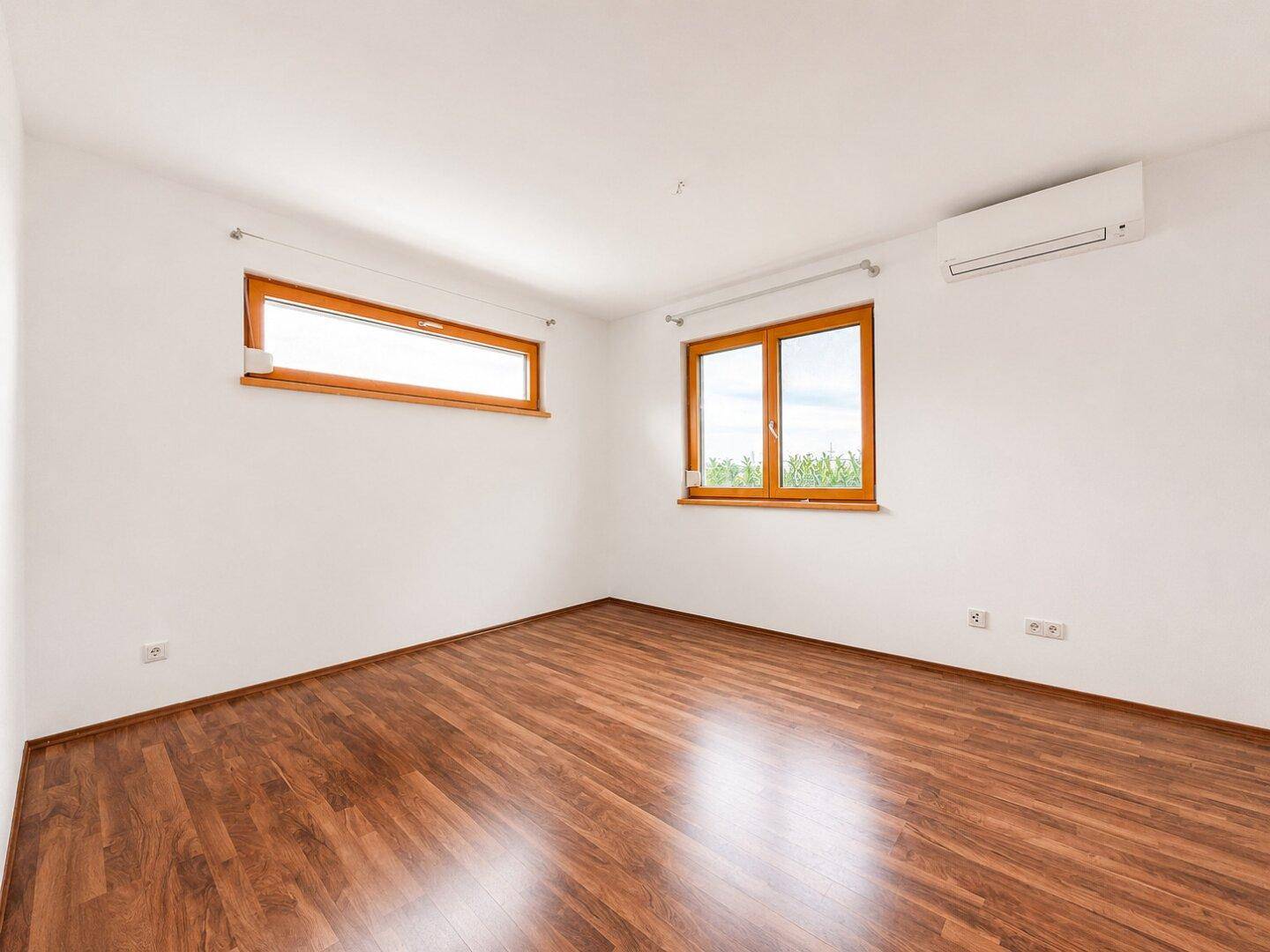 Leeres Zimmer mit dunklem Holzboden, weißen Wänden und Klimaanlage.