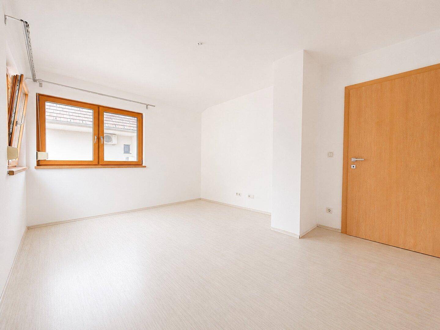Helles, leeres Zimmer mit hellem Holzboden, weißen Wänden und Klimaanlage.