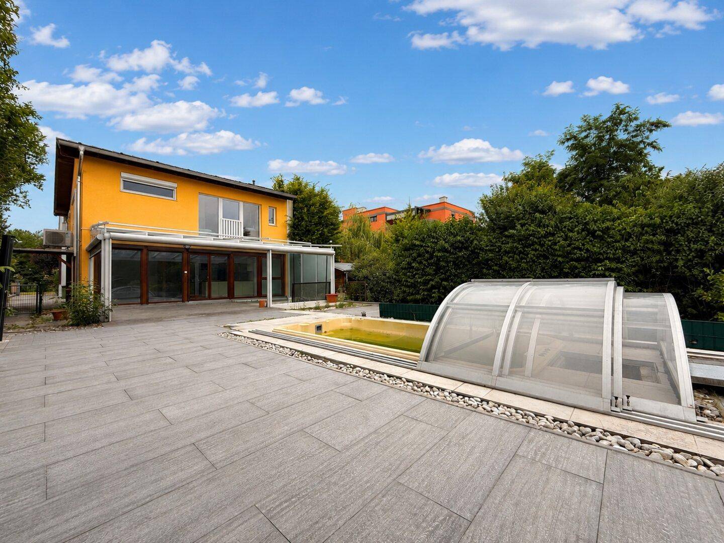 Geräumige geflieste Terrasse mit Pool und Schiebeabdeckung neben einem Haus mit gelber Fassade.