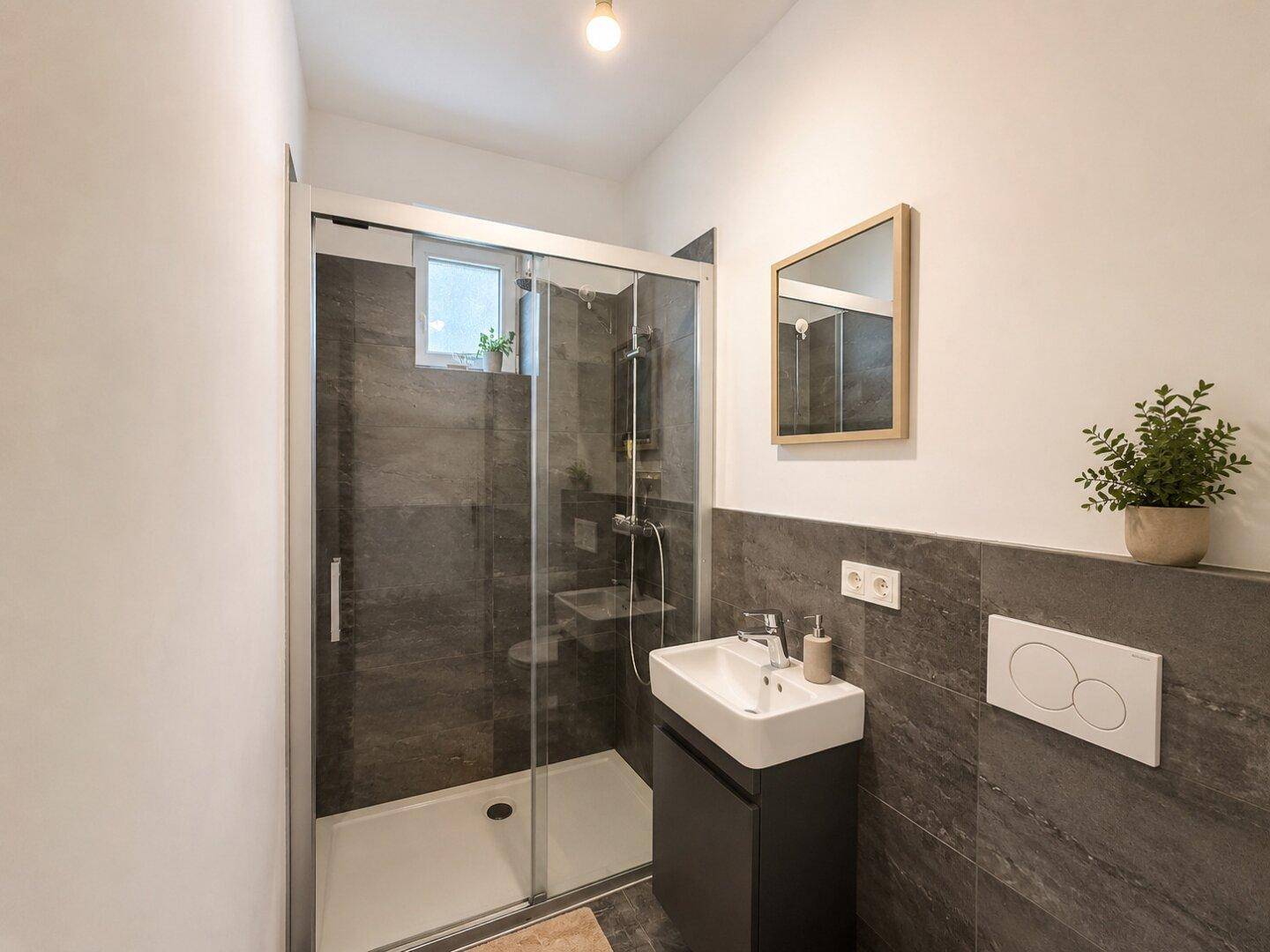 Modernes Badezimmer mit dunklen Fliesen, Duschkabine und einem kleinen Fenster für natürliche Belüftung.