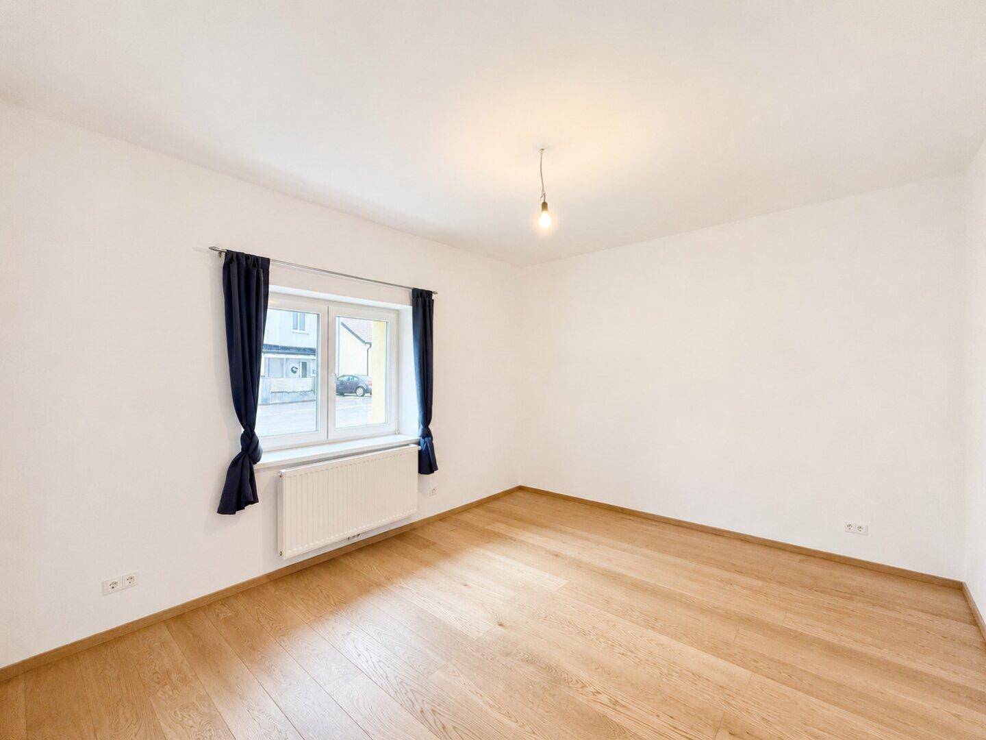 Geräumiges, leeres Wohnzimmer mit Holzboden, weißen Wänden und einem Fenster mit Blick auf die Straße.
