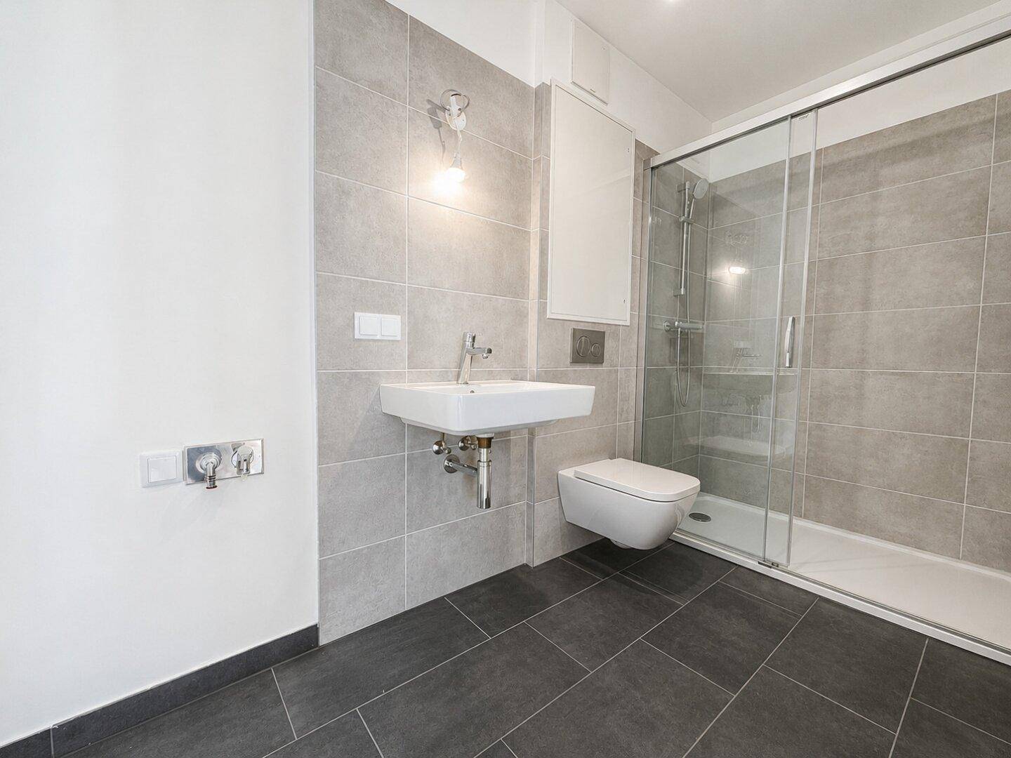 Modernes Badezimmer mit grauen Fliesen, wandhängendem WC, Waschbecken und einer begehbaren Dusche.
