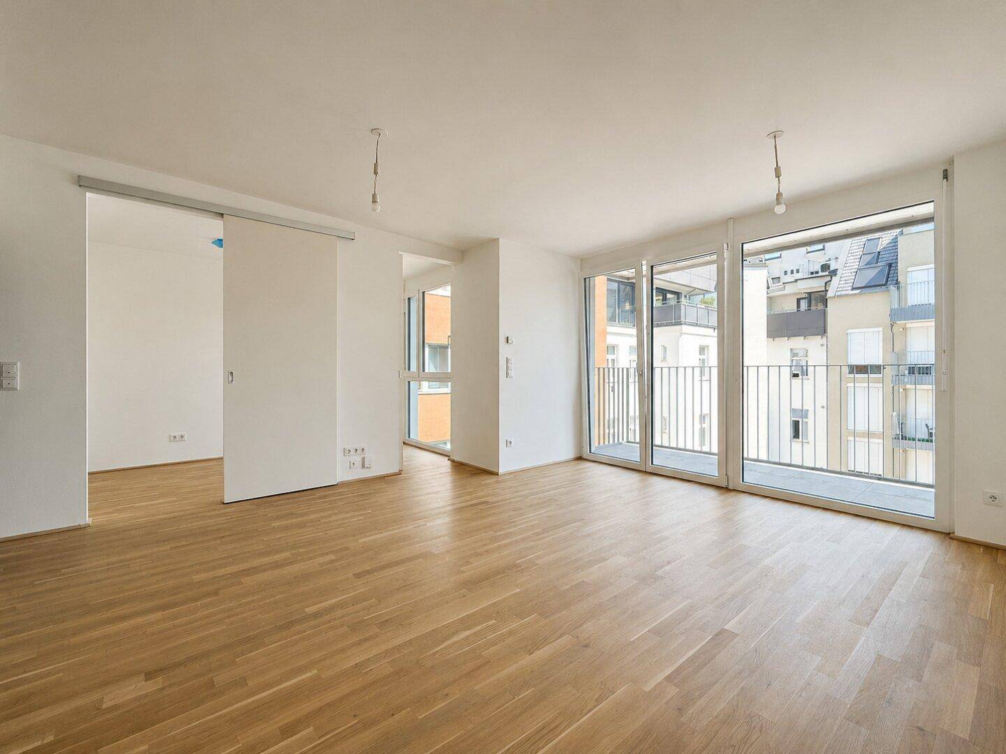Geräumiger, unmöblierter Wohnbereich mit hellem Parkettboden und großen Schiebetüren zum Balkon.