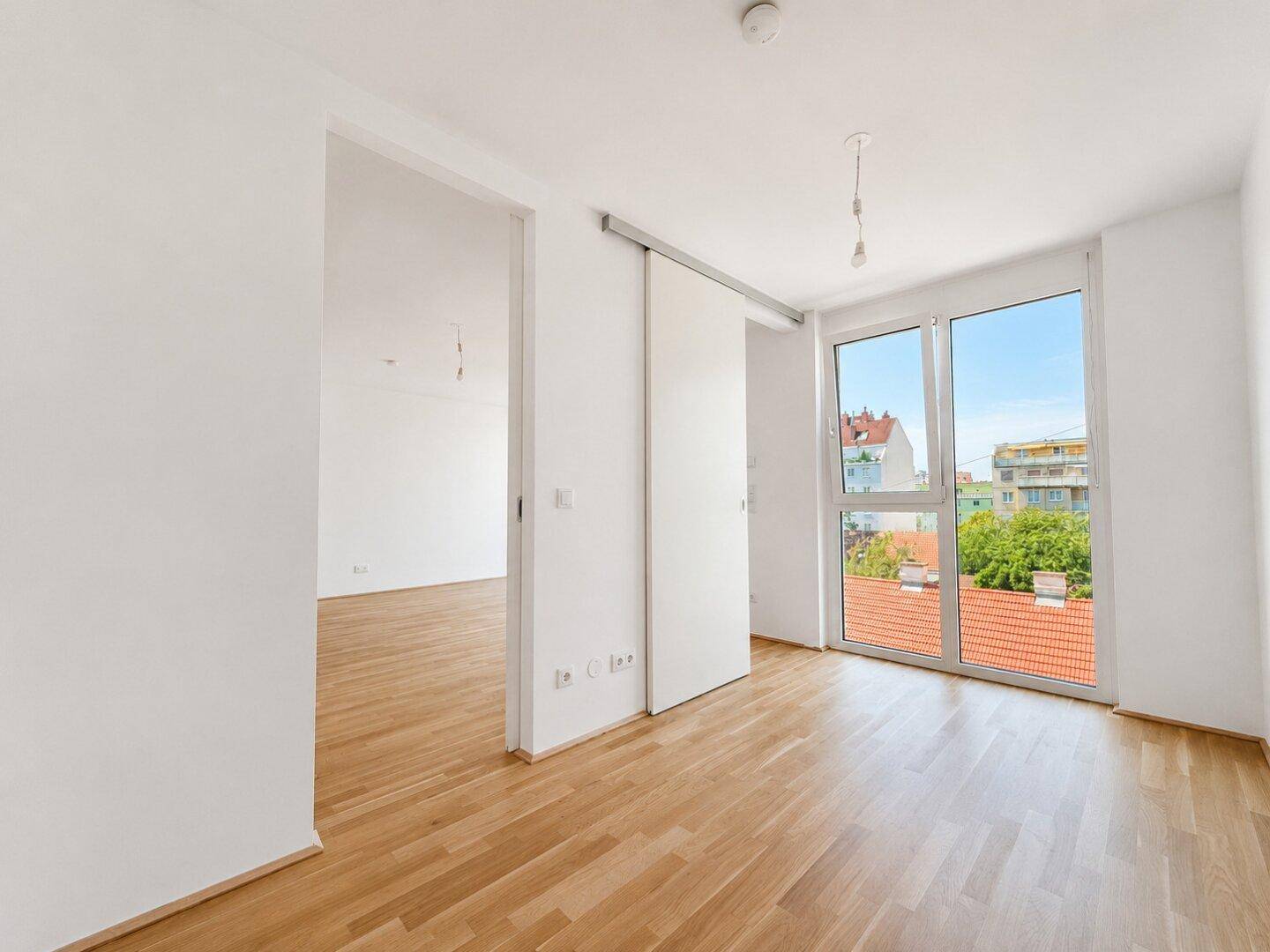 Helles, leeres Zimmer mit Holzboden, großen Fenstern und Schiebetür.