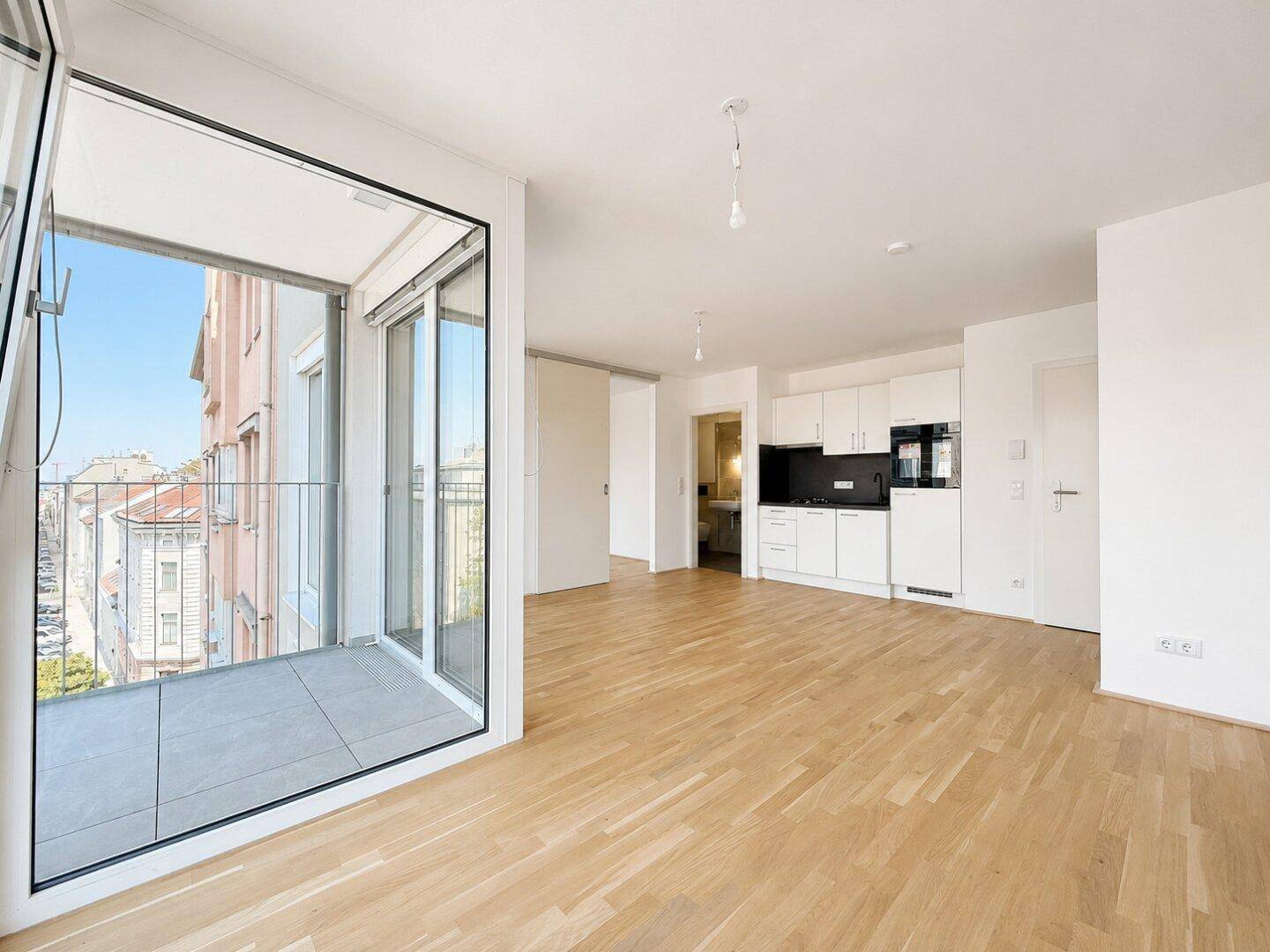 Heller, leerer Wohnbereich mit Holzboden, Balkon und vorbereiteter Küchenzeile.