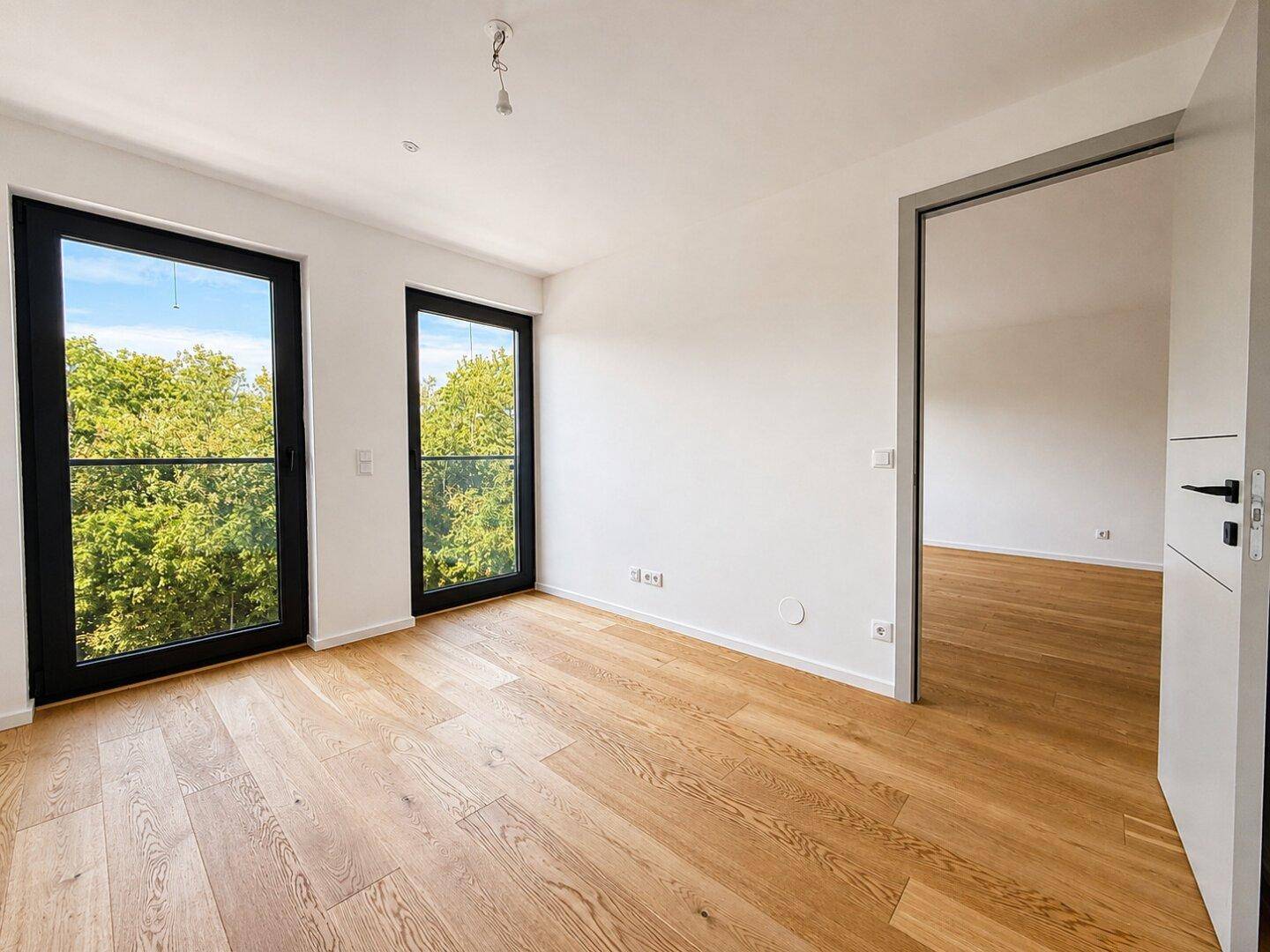 Leeres Schlafzimmer mit zwei hohen Fenstertüren und hellem Holzparkettboden.