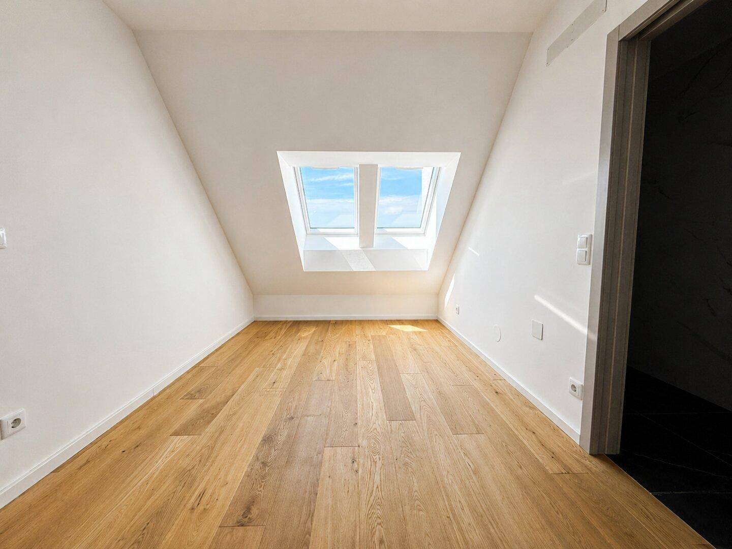 Helles, leeres Zimmer mit Holzboden und zwei Dachfenstern, bereit zur individuellen Gestaltung.