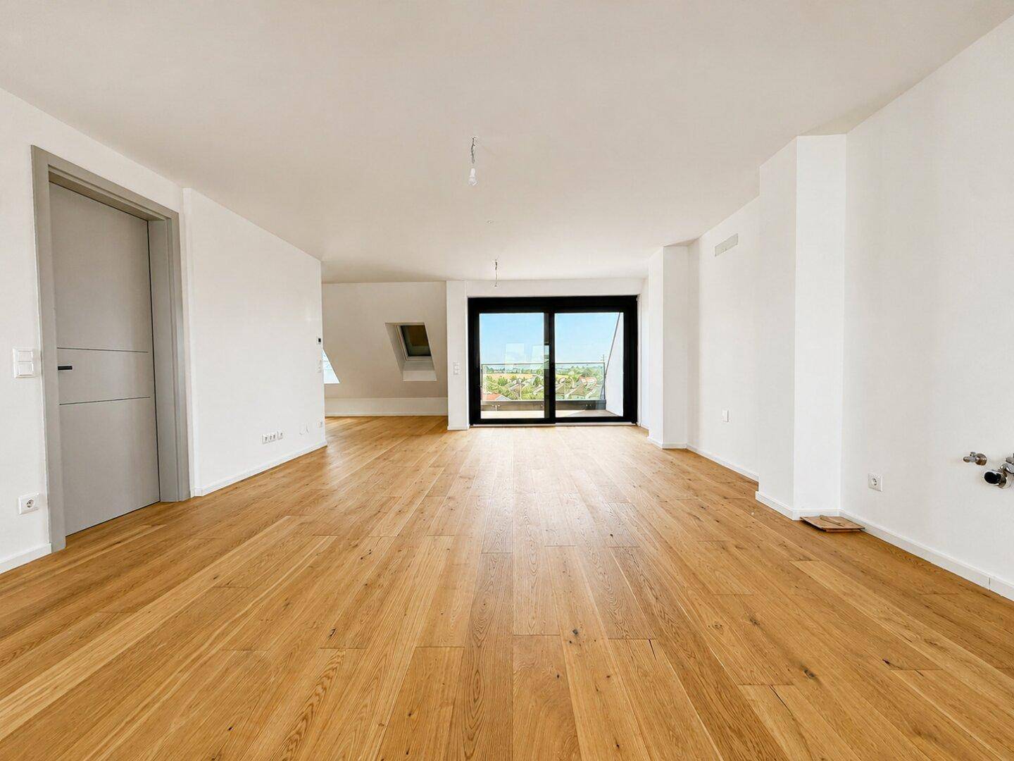 Geräumiger, leerer Wohnbereich mit hellem Holzboden und Zugang zu einem Balkon oder einer Terrasse.