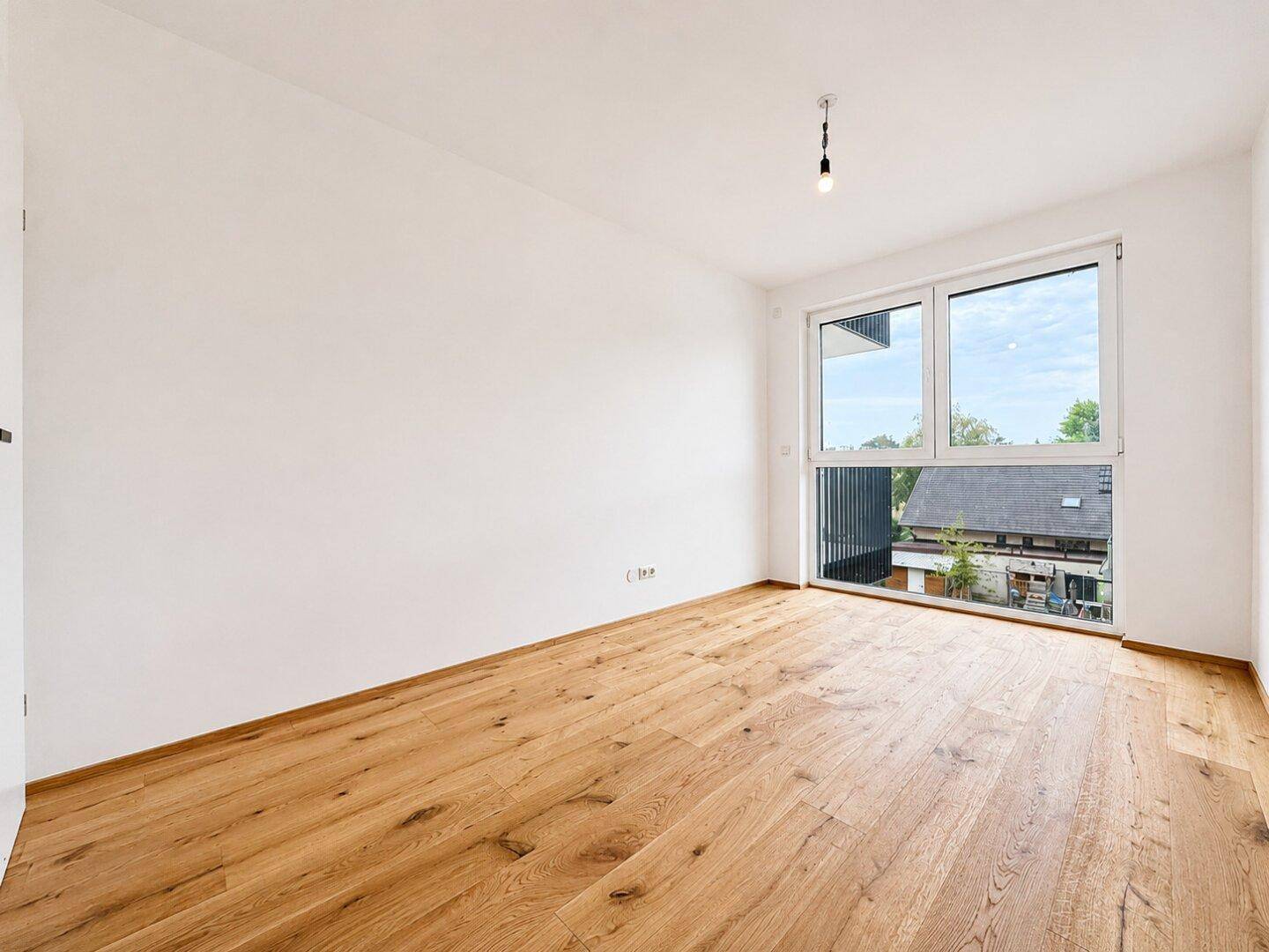 Helles Zimmer mit Holzboden und großem Fenster, bietet viel Tageslicht und eine angenehme Atmosphäre.