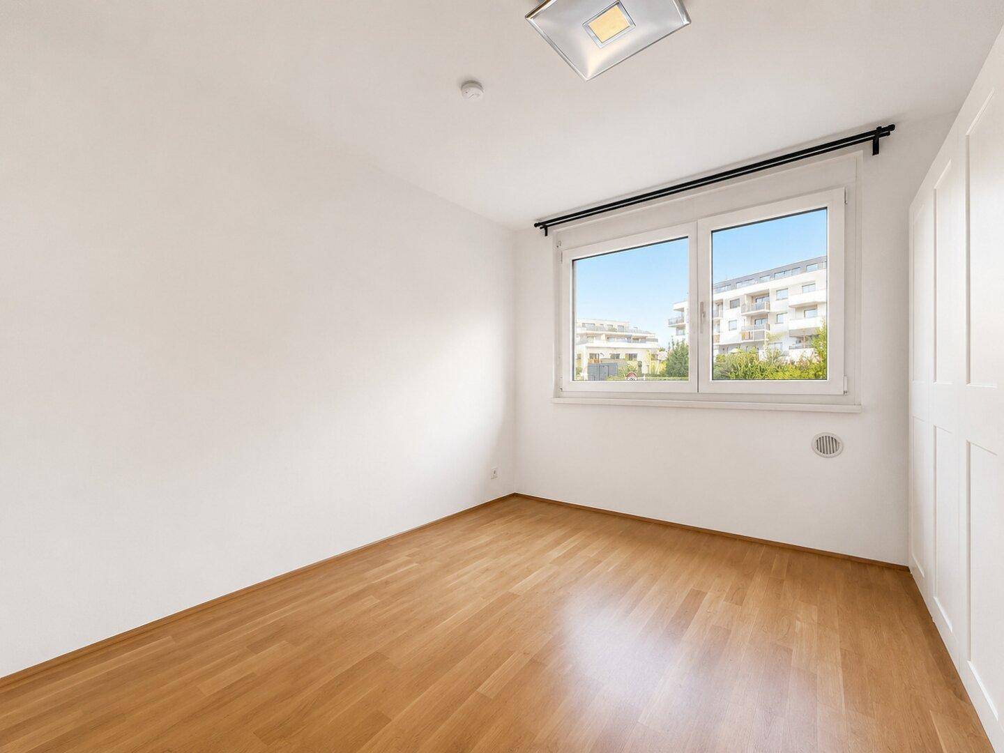 Helles, leeres Schlafzimmer mit Holzboden und einem Fenster mit Blick auf umliegende Gebäude.