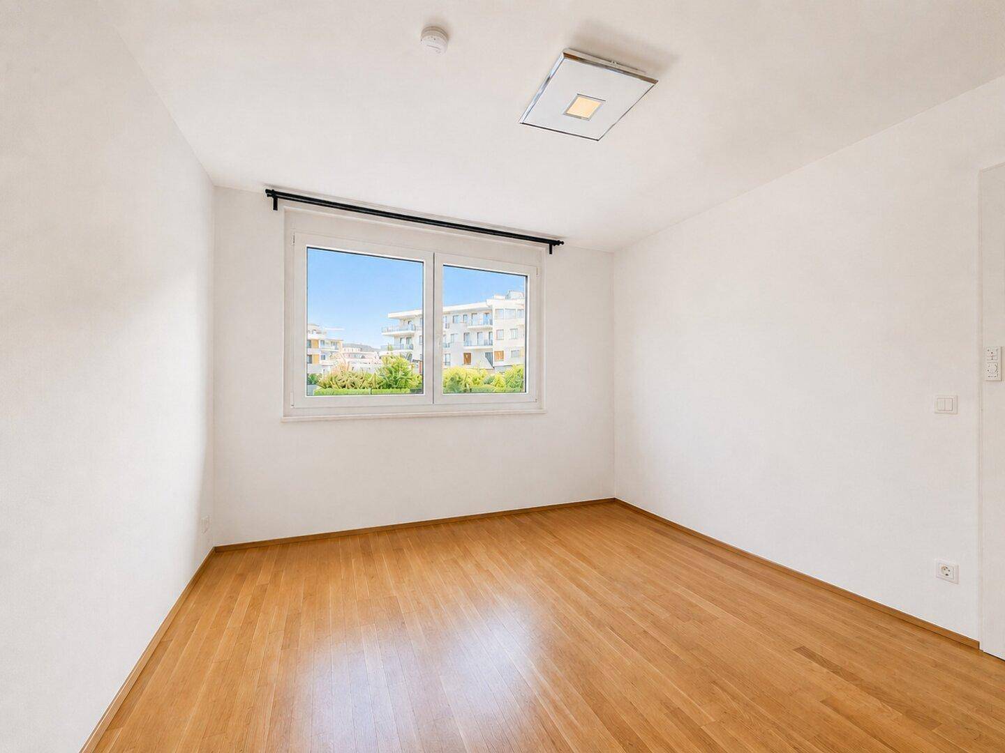 Geräumiges, leeres Schlafzimmer mit Holzboden und großem Fenster, das viel Tageslicht hereinlässt.