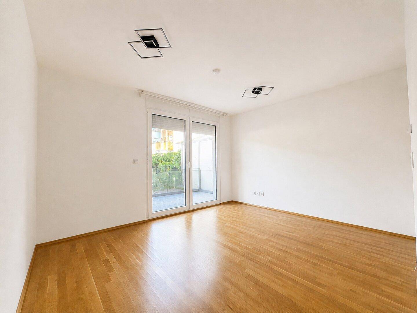 Leeres, helles Wohnzimmer mit Holzboden und großen Glastüren zum Balkon oder zur Terrasse.