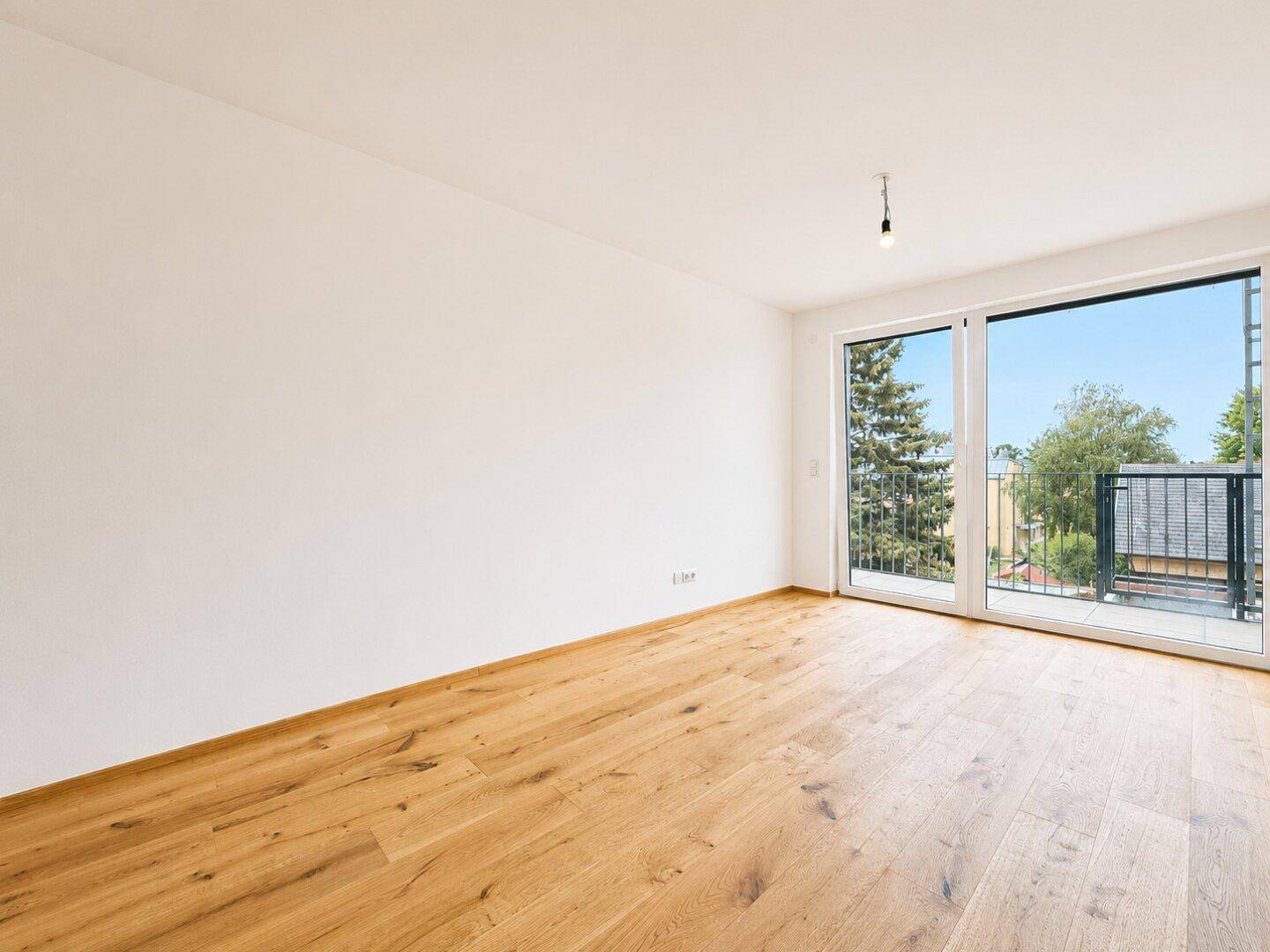 Leeres Zimmer mit hellem Holzboden und großen Fenstern, die viel Tageslicht hereinlassen.