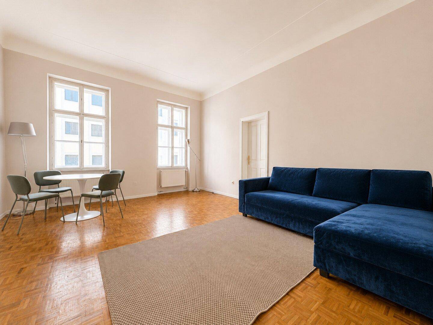 Geräumiges Wohnzimmer mit blauem Ecksofa, Esstisch und klassischem Parkettboden, hell und einladend.