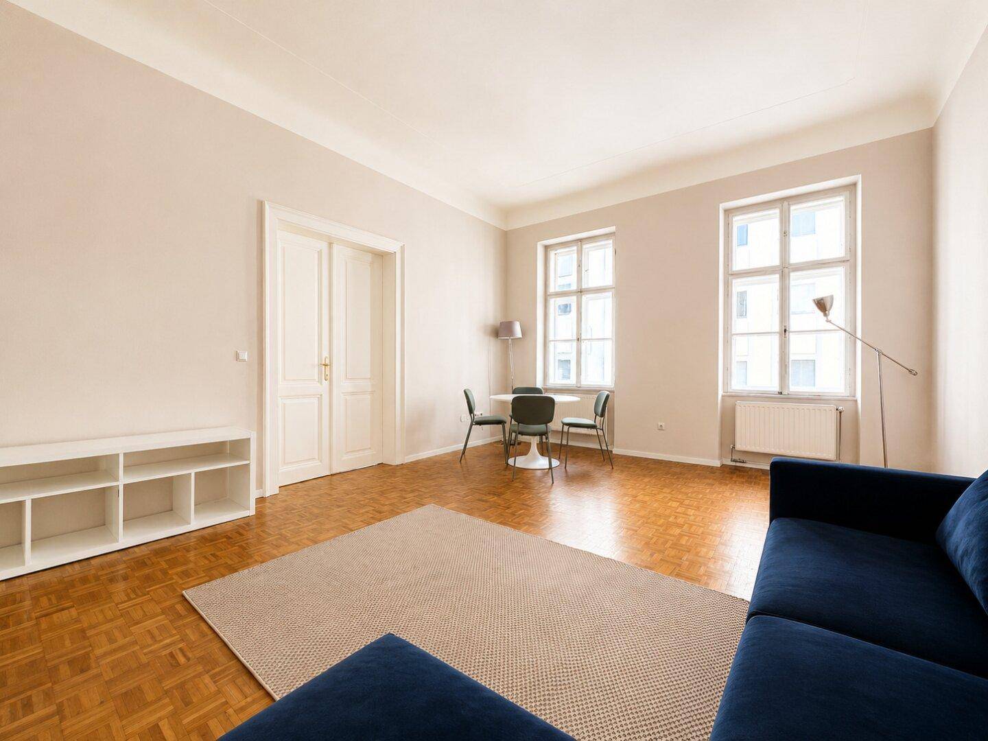 Weitläufiges Wohnzimmer mit Parkettboden, blauer Couch und weißem Regal, durch Doppeltüren zugänglich.