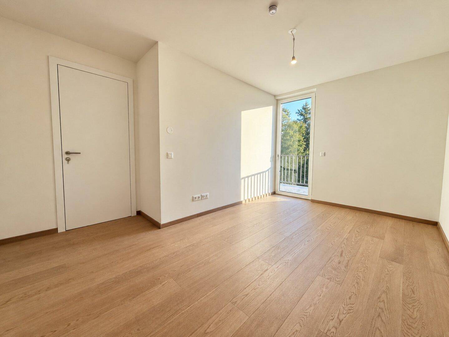 Helles, leeres Schlafzimmer mit Holzboden und Zugang zu einem kleinen Balkon.