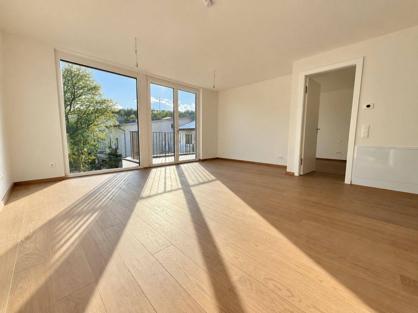 Geräumiges, leeres Wohnzimmer mit großen Fenstern, Holzboden und viel natürlichem Licht.
