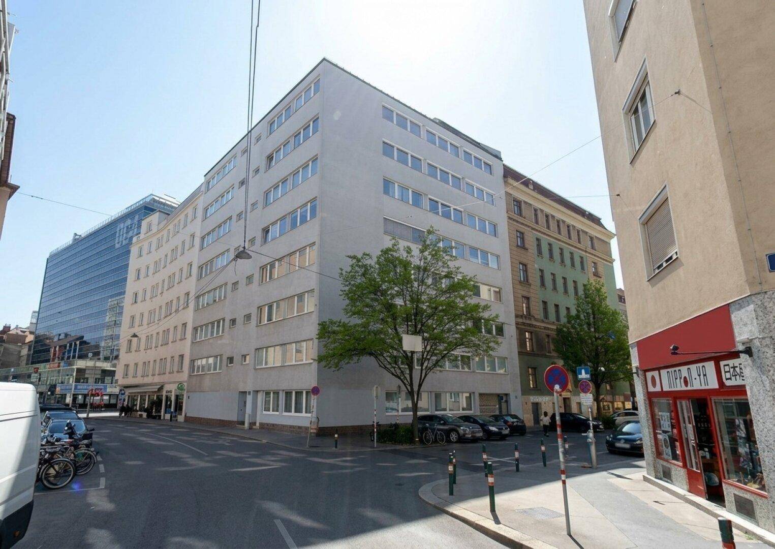 Modernes Mehrfamilienhaus an einer Straßenecke mit vielen Fenstern und Bäumen im städtischen Umfeld.